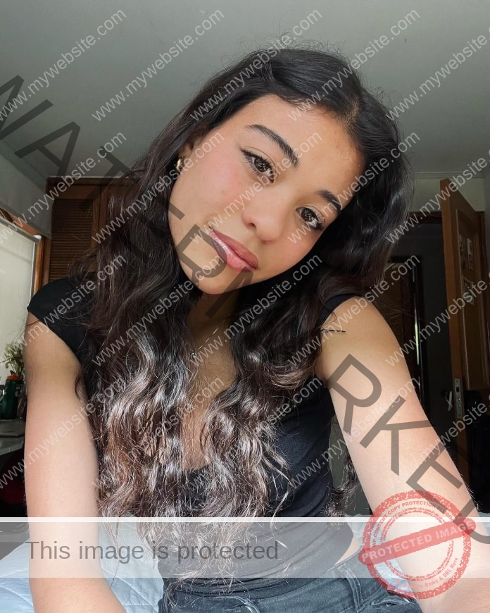 Ampara Herrera, a young woman with wavy dark hair and a black top, smiles on a sunlit bed; door and window show daily Chilean life.