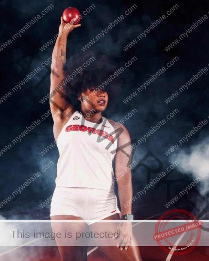 Ana Da Silva, a female athlete in a white GEORGIA uniform, holds a shot put above her head on a misty track, looking confident.