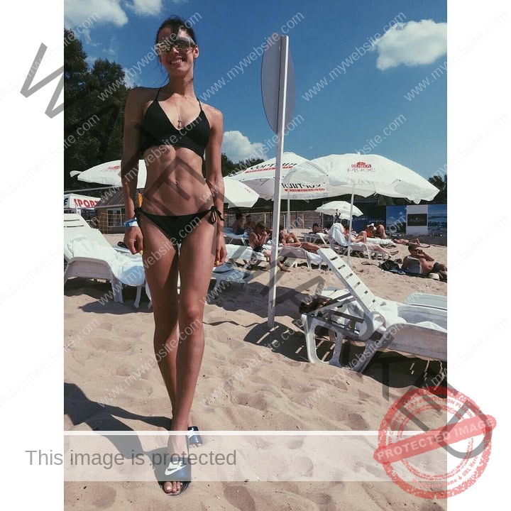 anastasia-bryzgina-ukraine-bryzgina_a-03831 Anastasia Bryzgina, track star from Ukraine, in a black bikini and sunglasses stands on a sunny beach near white lounge chairs.