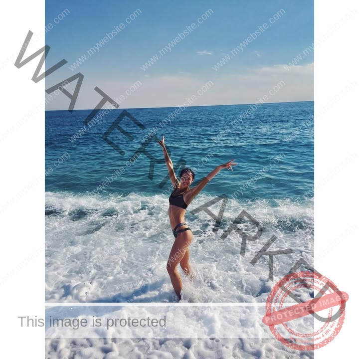 anastasia-bryzgina-ukraine-bryzgina_a-03896 Anastasia Bryzgina, track star from Ukraine, poses joyfully in a bikini with arms raised in shallow ocean waves under sunny blue skies.