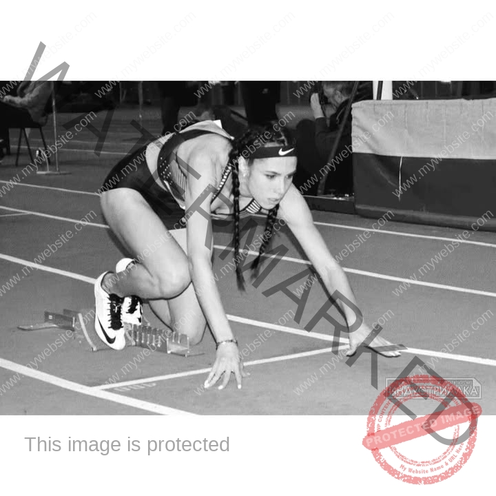 anastasia-bryzgina-ukraine-bryzgina_a-03902 Anastasia Bryzgina, track star from Ukraine, with braided hair, crouches at starting blocks on an indoor track, focused ahead. Black and white.