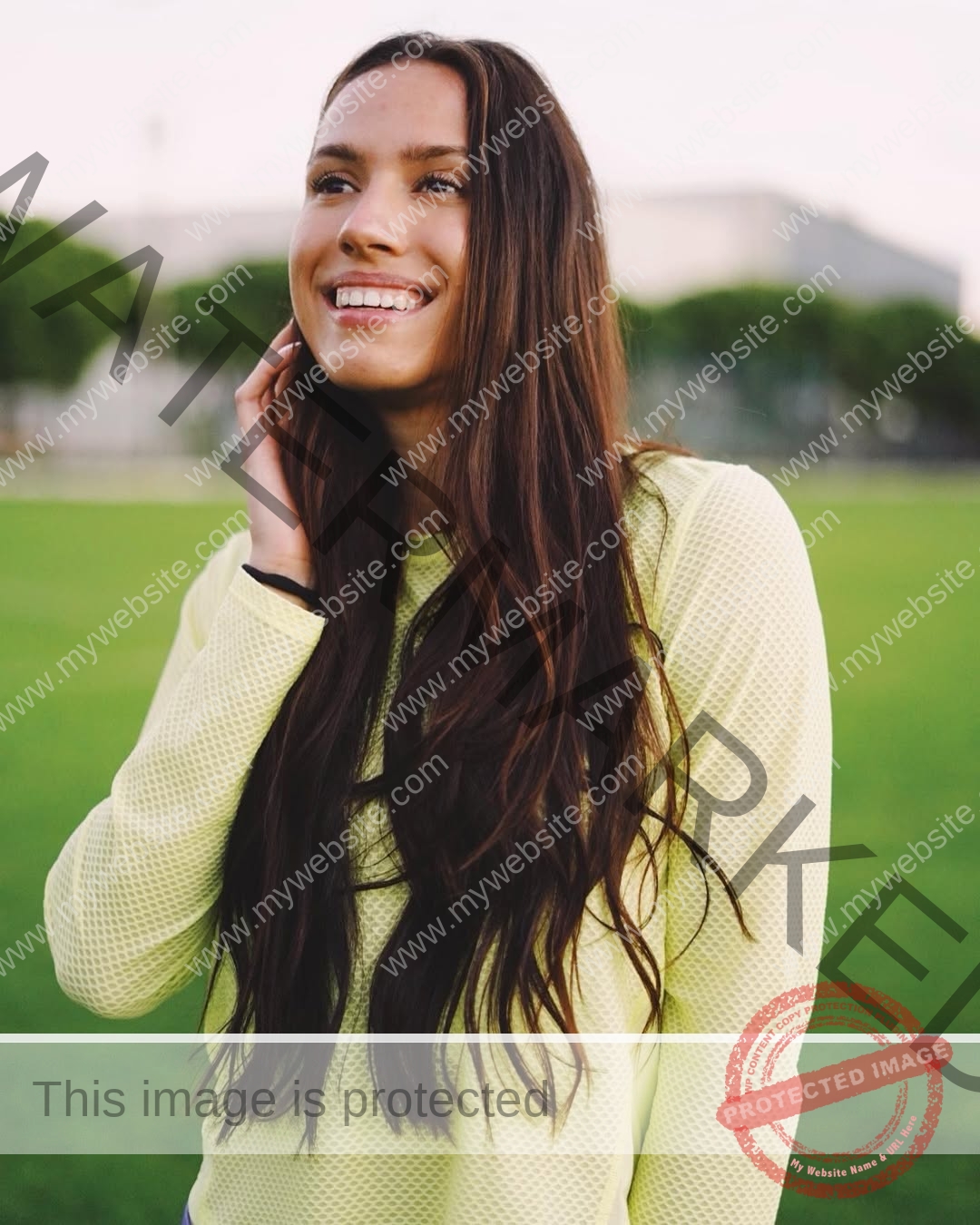 anastasia-bryzgina-ukraine-bryzgina_a-03972 Anastasia Bryzgina, track star from Ukraine, with long brown hair in a yellow shirt, smiles on a grassy field, trees blurred behind.