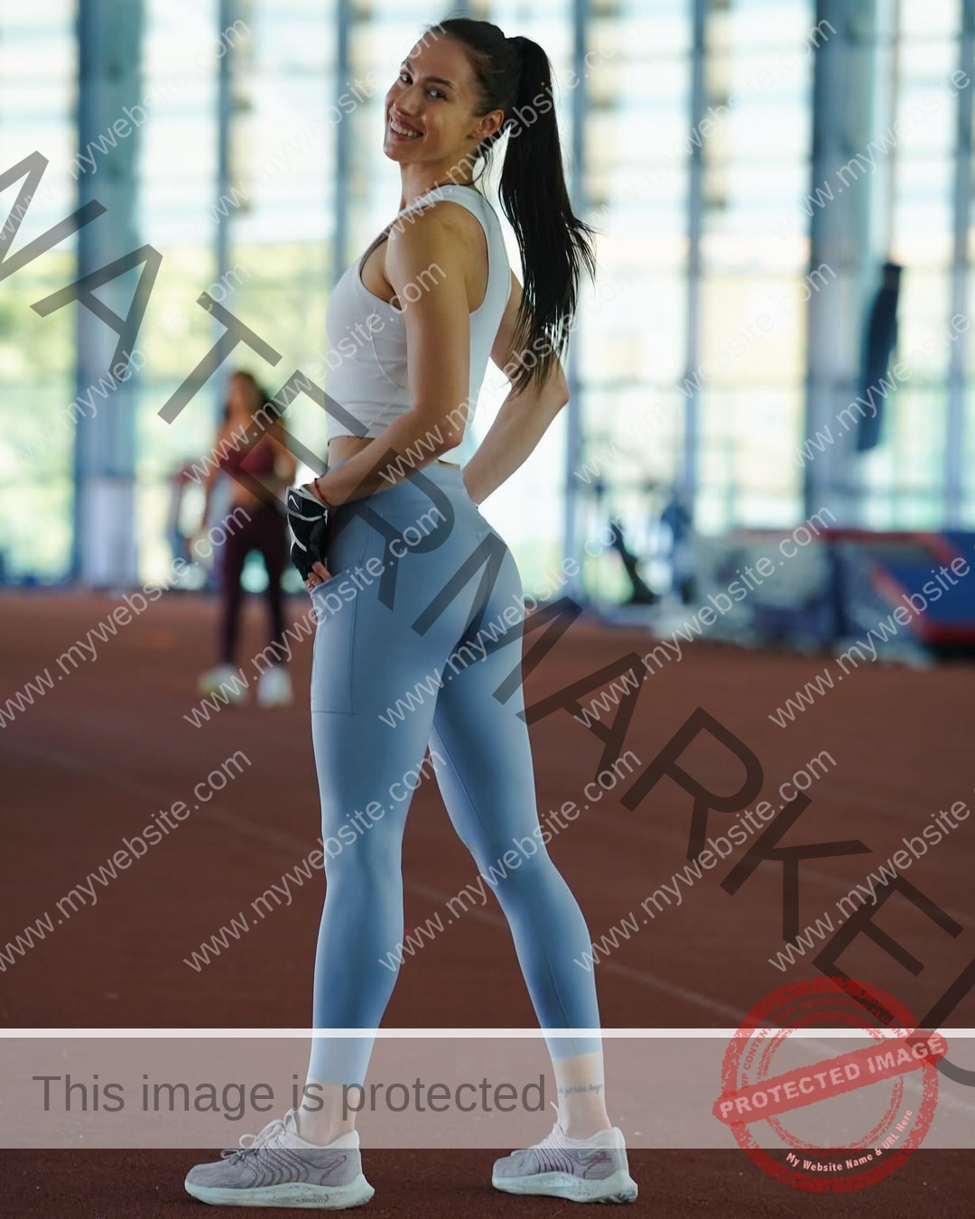 anastasia-bryzgina-ukraine-bryzgina_a-04070 Anastasia Bryzgina, track star from Ukraine, in a white crop top and blue leggings smiles on an indoor track with gear behind her.