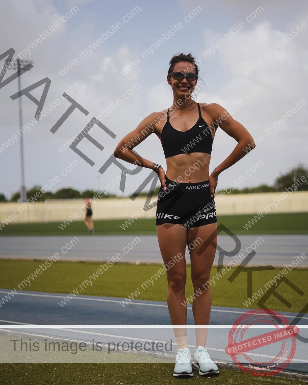 anastasia-bryzgina-ukraine-bryzgina_a-04078 Anastasia Bryzgina, track star from Ukraine, smiles in athletic wear on a running track with hands on hips; another person stands behind.