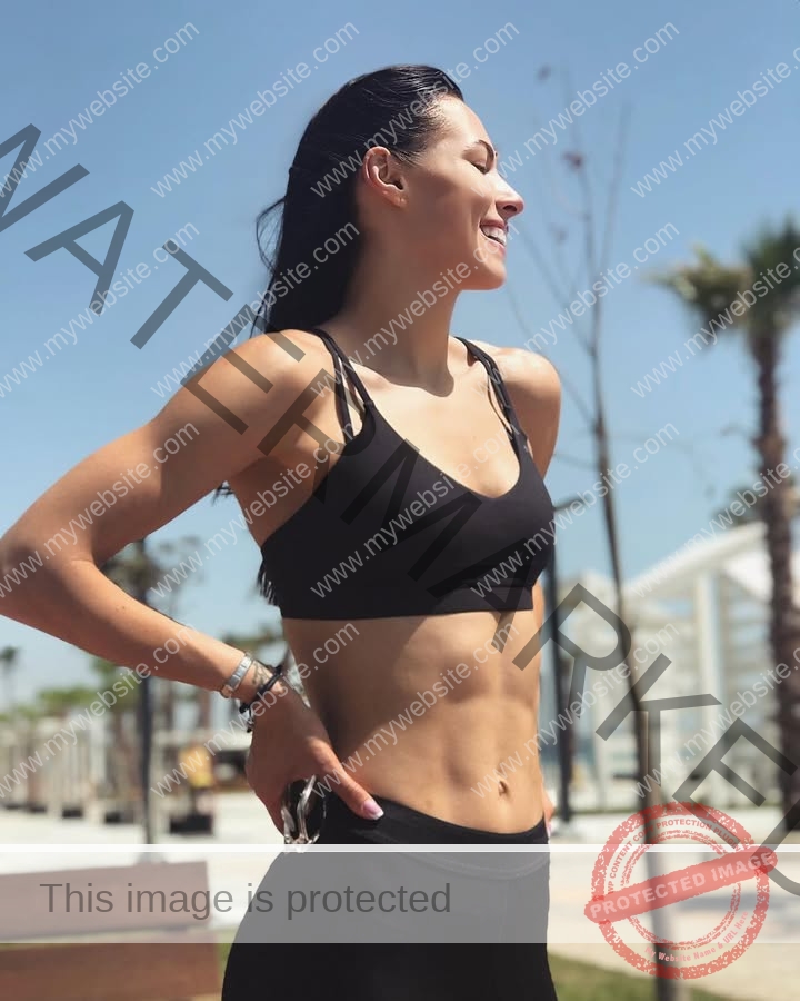 anastasia-bryzgina-ukraine-bryzgina_a-04130 Anastasia Bryzgina, track star from Ukraine, stands outside in black workout gear, smiling with defined abs; palms and a bench behind.
