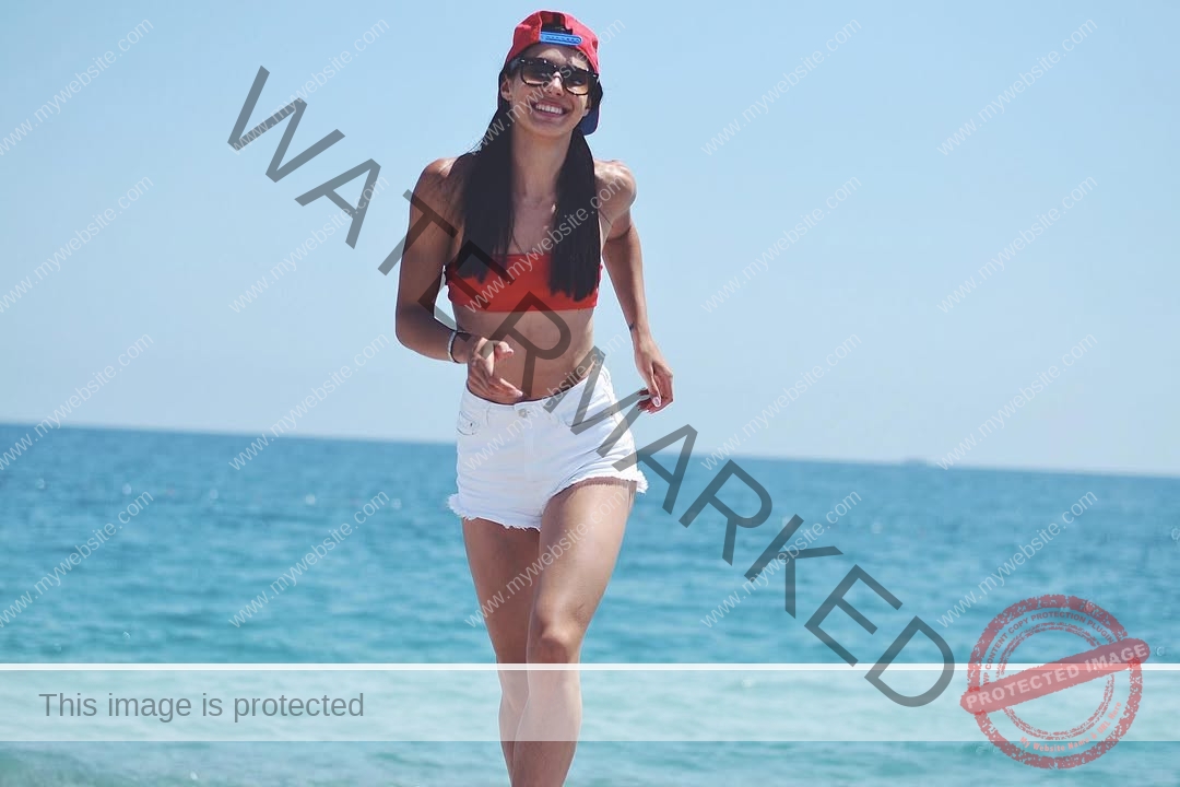 anastasia-bryzgina-ukraine-bryzgina_a-04136 Anastasia Bryzgina, track star from Ukraine, smiles in a red bikini top, white shorts, cap, and sunglasses on a sunny beach.