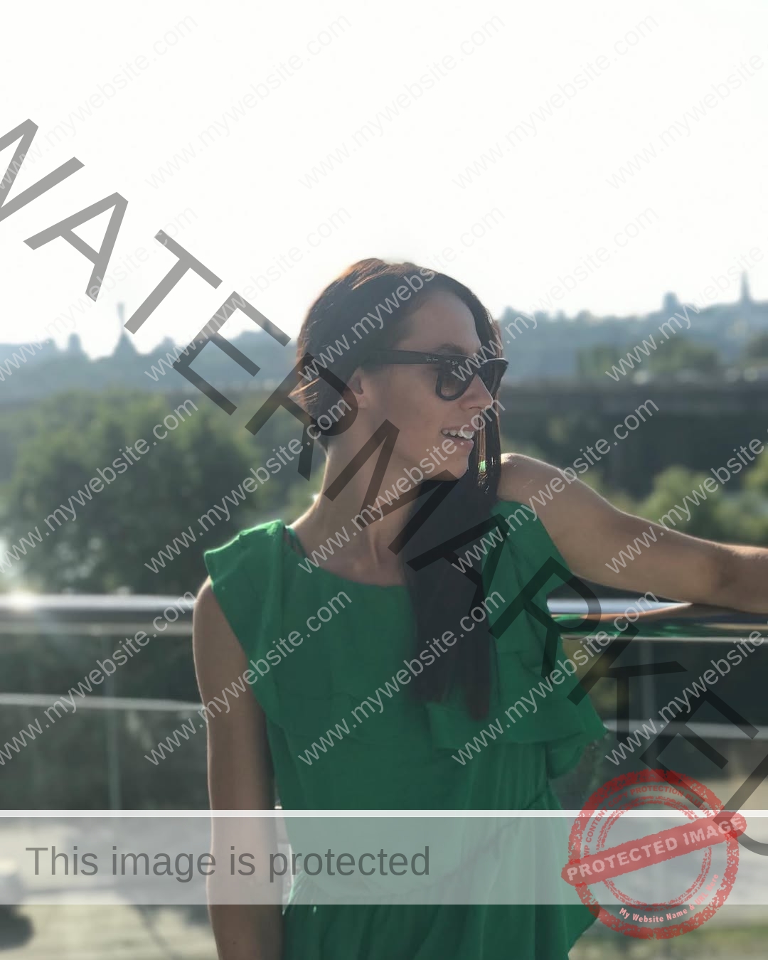 anastasia-bryzgina-ukraine-bryzgina_a-04148 Anastasia Bryzgina, track star from Ukraine, stands by a railing in a green dress and sunglasses, smiling outdoors on a sunny day.