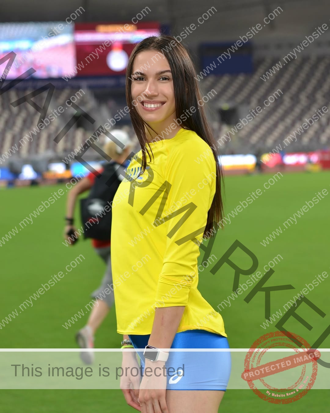 anastasia-bryzgina-ukraine-bryzgina_a-04154 Anastasia Bryzgina, track star from Ukraine, with long brown hair in a yellow shirt and blue shorts, smiles on a green sports field.