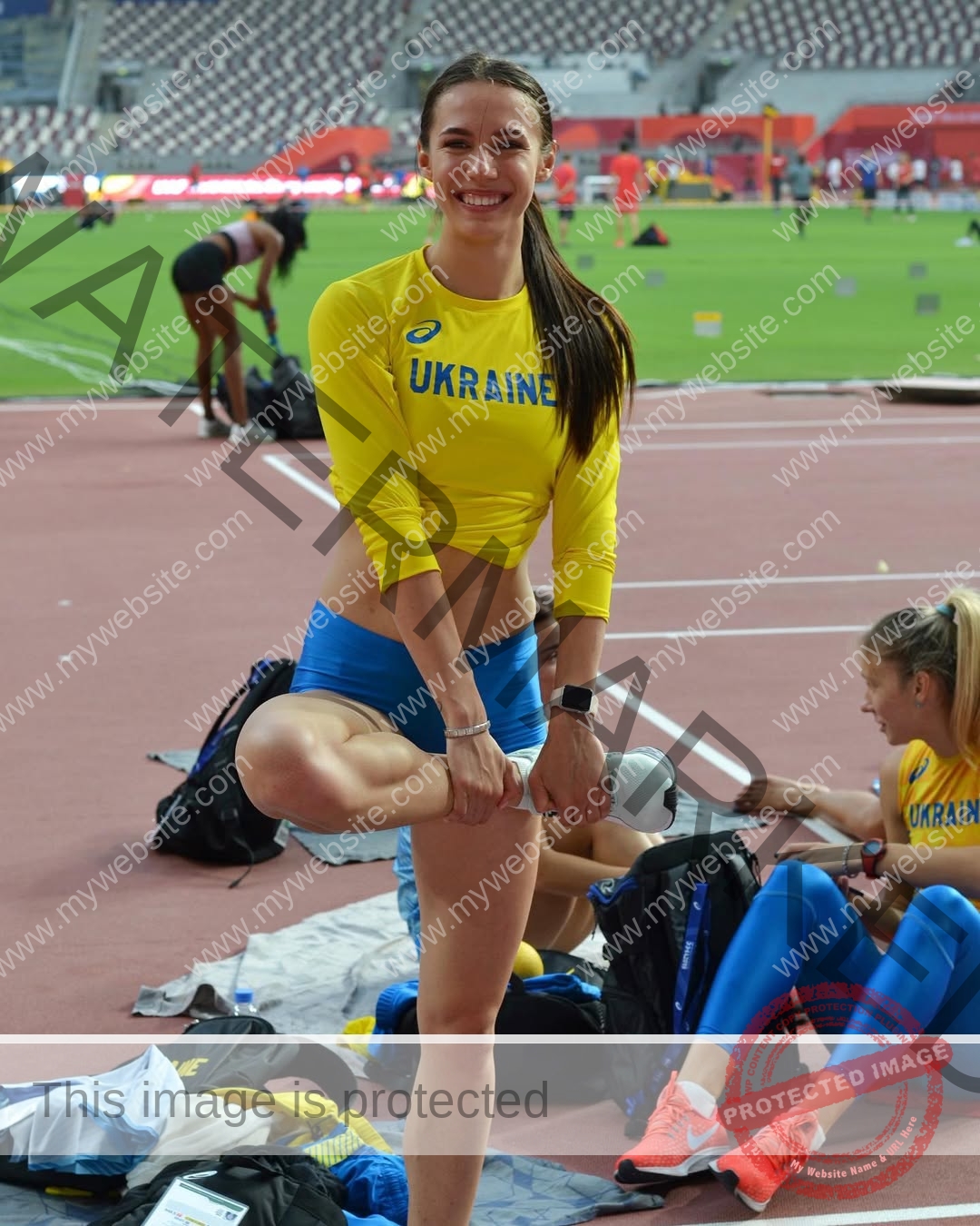 anastasia-bryzgina-ukraine-bryzgina_a-04156 Anastasia Bryzgina, track star from Ukraine, stretches her leg on a track in yellow and blue as teammates and stadium seats appear nearby.