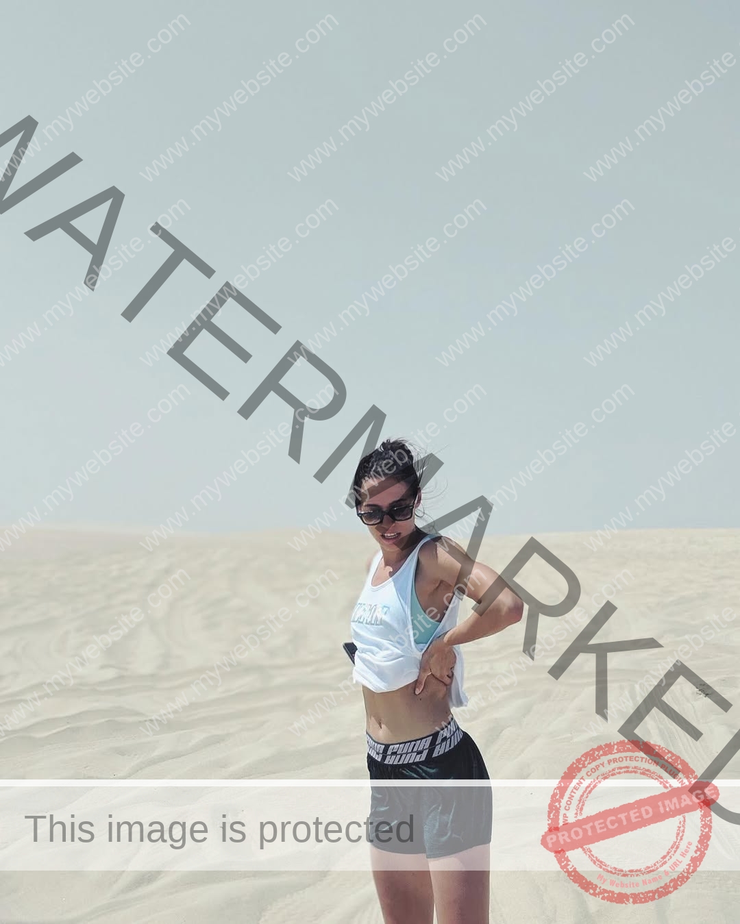 anastasia-bryzgina-ukraine-bryzgina_a-04158 Anastasia Bryzgina, track star from Ukraine, wearing a white crop top, black shorts, and sunglasses stands on a sandy desert.