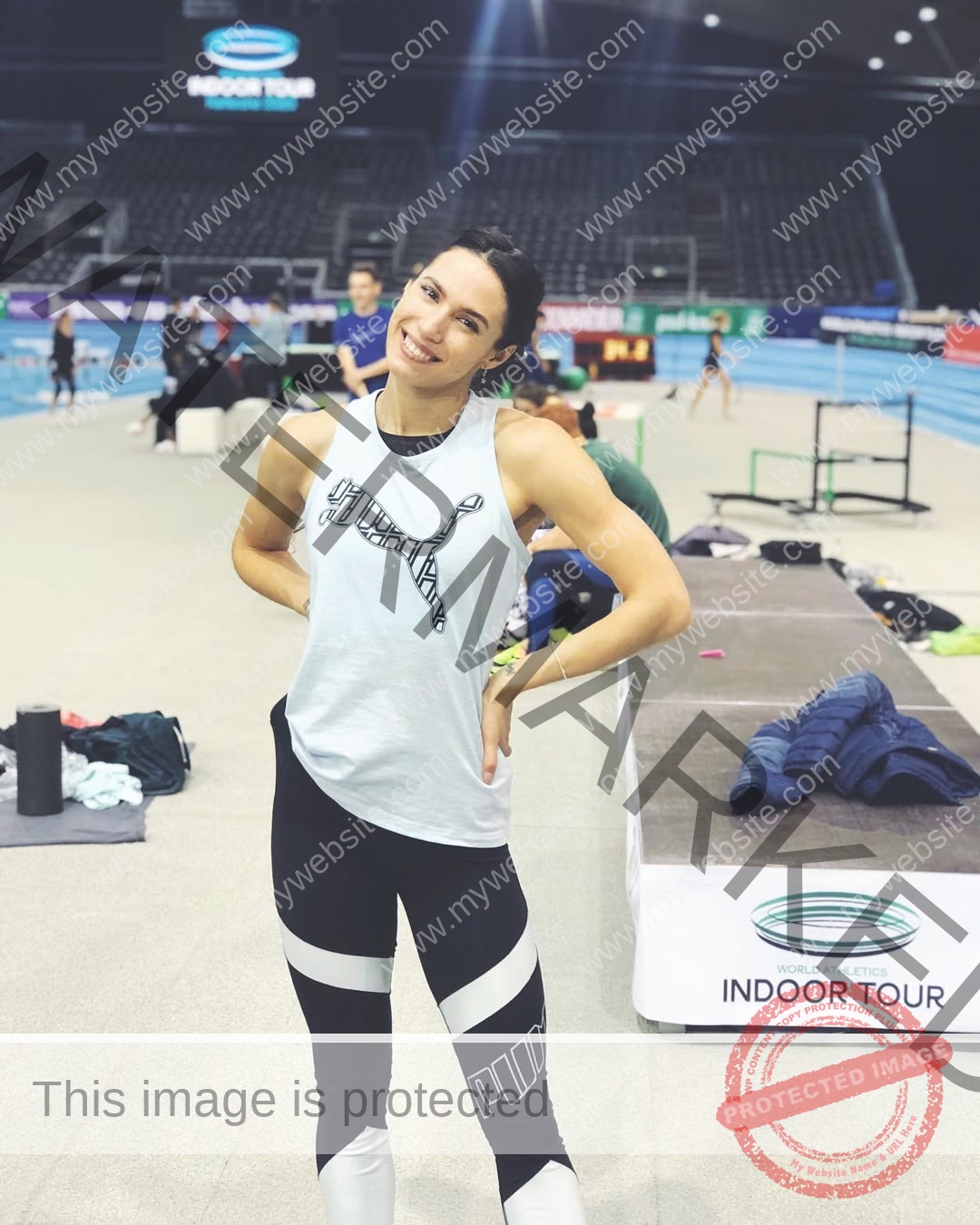 anastasia-bryzgina-ukraine-bryzgina_a-04185 Anastasia Bryzgina, track star from Ukraine, stands smiling on an indoor track, hands on hips; sports gear and event sign behind.