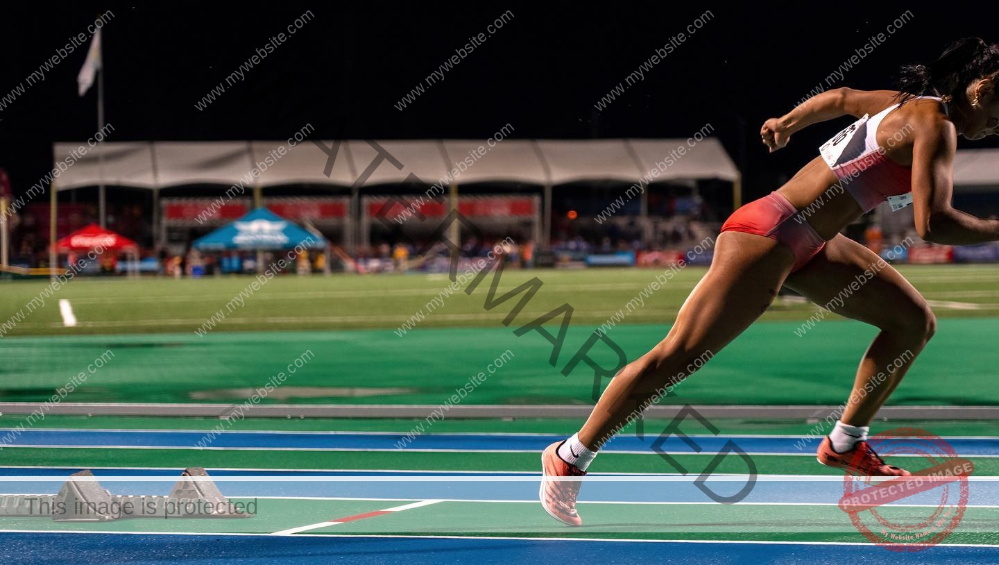 andrea-rivera-aviles-puerto-rico-aandrea.sofiaa-05886 Andrea Rivera Aviles, track athlete from Puerto Rico, launches off starting blocks in red gear on a blue track at night with blurred crowd.