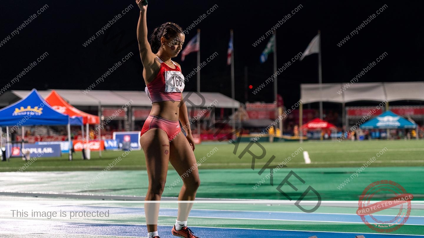 andrea-rivera-aviles-puerto-rico-aandrea.sofiaa-05887 Andrea Rivera Aviles, track athlete from Puerto Rico, in red uniform and bib 166 stands on a track with one arm raised at night.