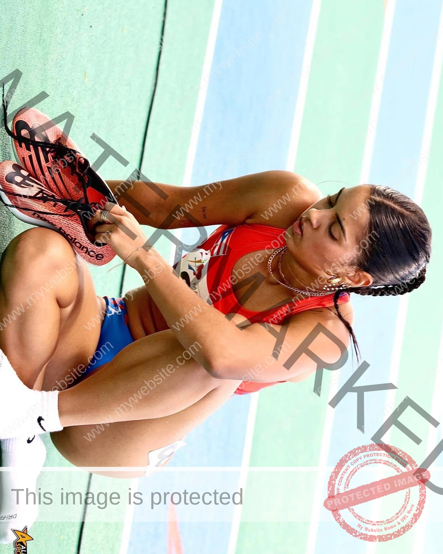 andrea-rivera-aviles-puerto-rico-aandrea.sofiaa-05899 Andrea Rivera Aviles, track athlete from Puerto Rico, sits on green track in red sports bra and blue shorts, tightening her spikes.