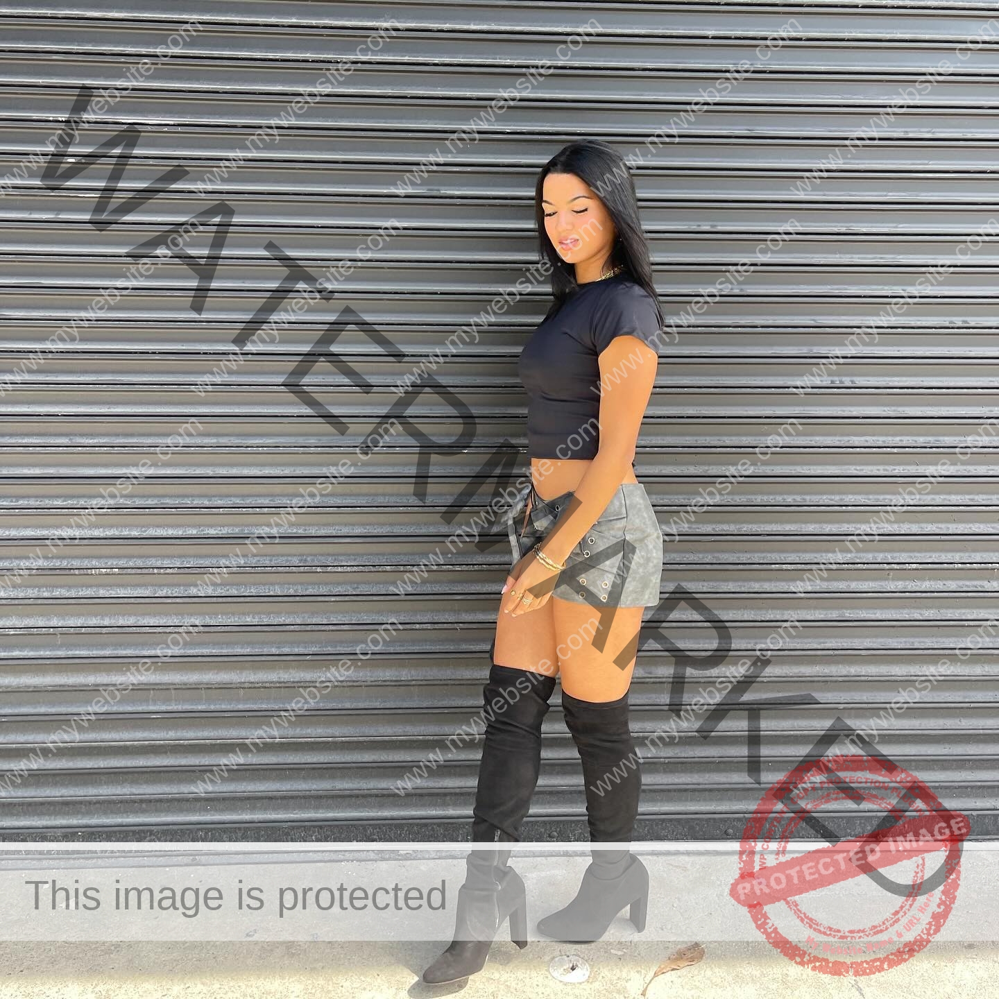 andrea-rivera-aviles-puerto-rico-aandrea.sofiaa-05903 Andrea Rivera Aviles, track athlete from Puerto Rico stands sideways by a gray shutter in a crop top, shorts, and knee-high boots.