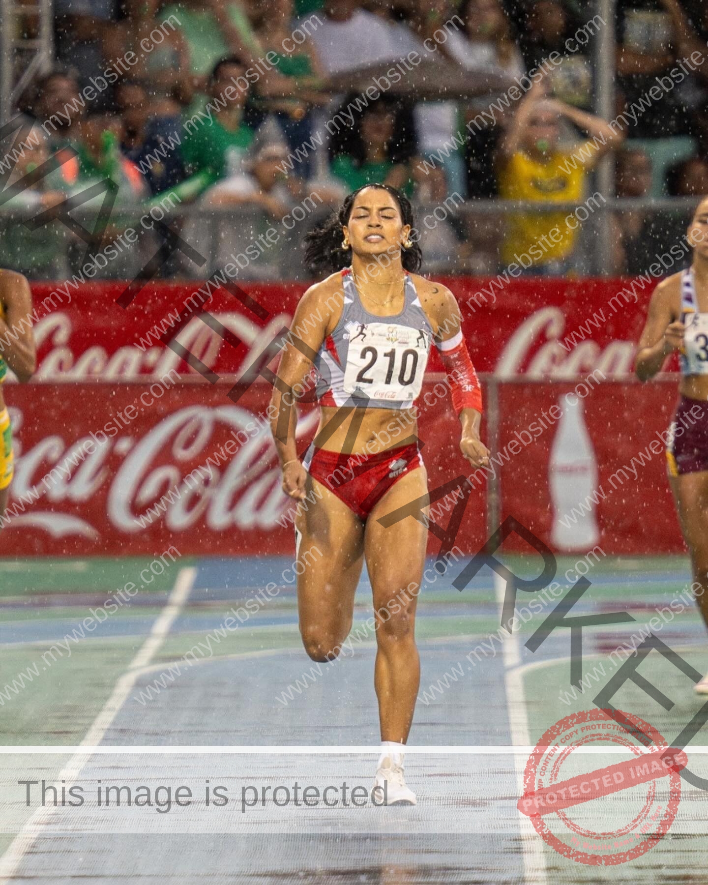 andrea-rivera-aviles-puerto-rico-aandrea.sofiaa-05905 Andrea Rivera Aviles, track athlete from Puerto Rico, in a red and white uniform, bib 210, sprints on a rainy track near the finish.