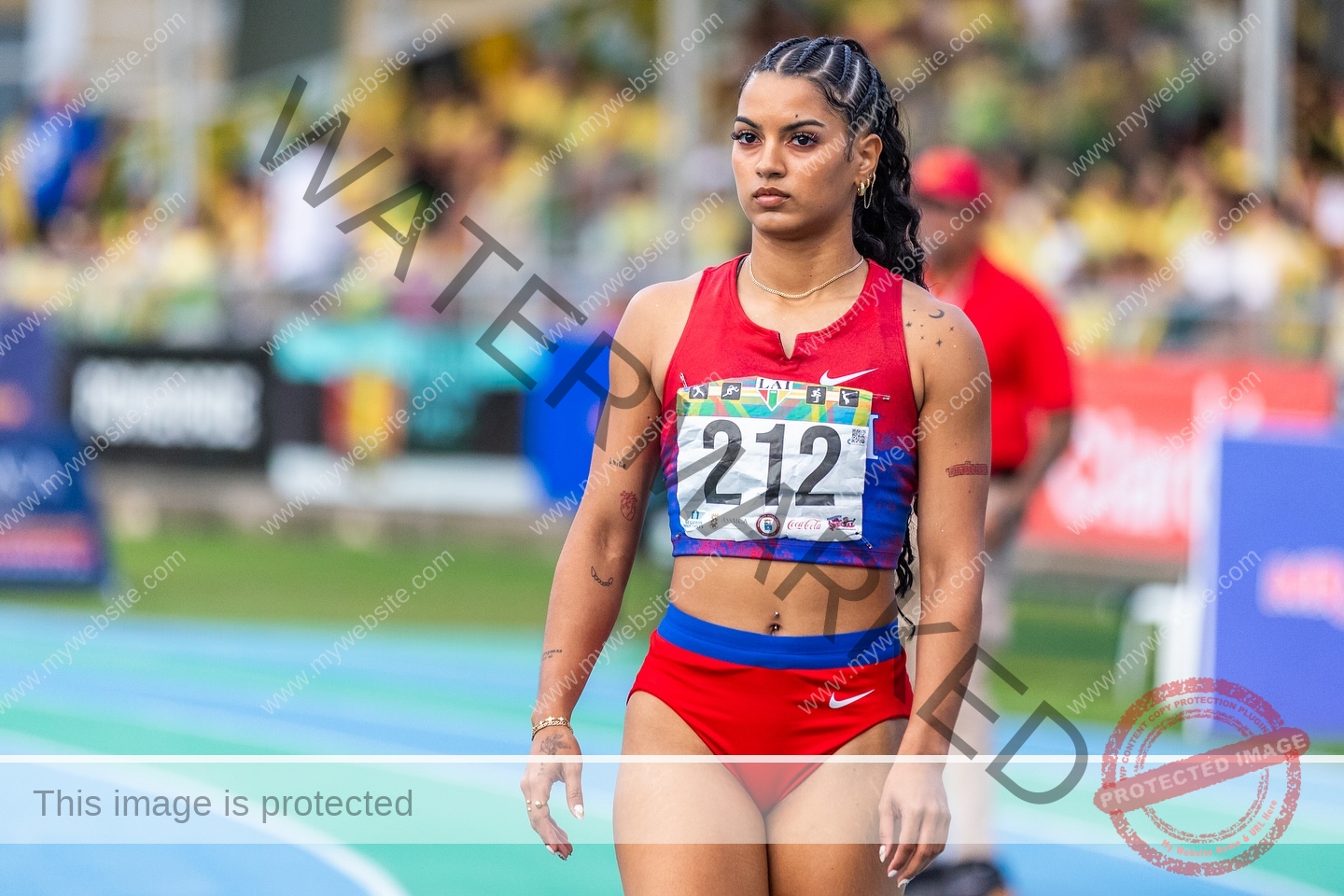 andrea-rivera-aviles-puerto-rico-aandrea.sofiaa-05939 Andrea Rivera Aviles, track athlete from Puerto Rico, in red sports bra and shorts (212), walks focused on the track, stadium behind.