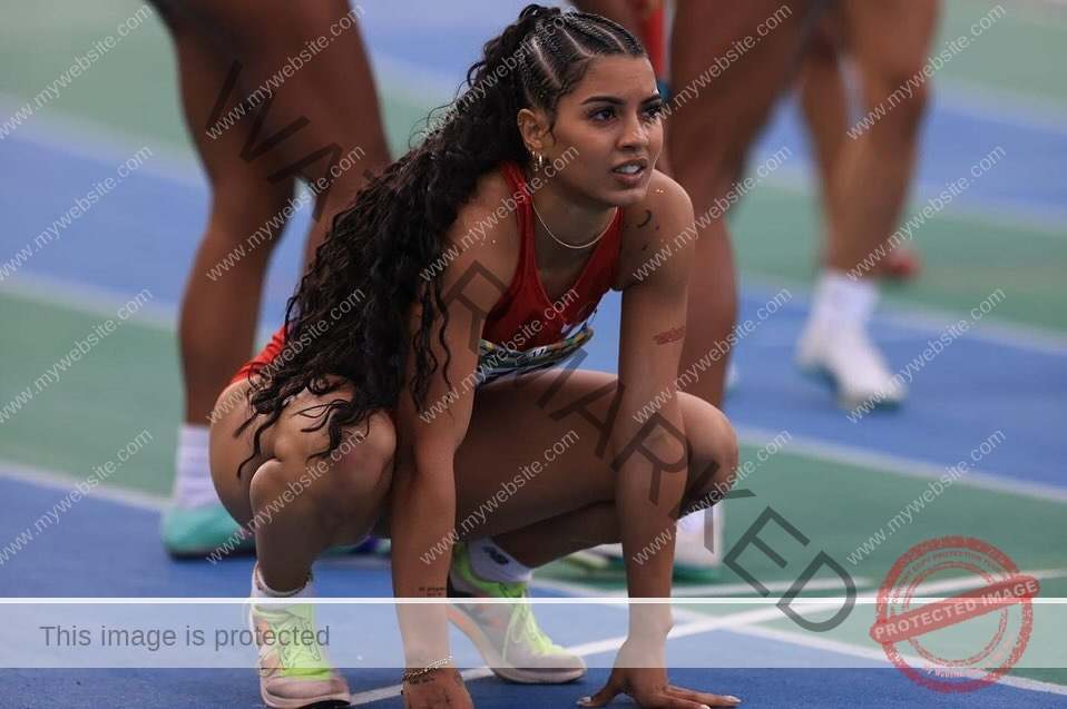 andrea-rivera-aviles-puerto-rico-aandrea.sofiaa-05947 Andrea Rivera Aviles, track athlete from Puerto Rico, with long curly hair in red uniform crouches at a blue and green track's start line.
