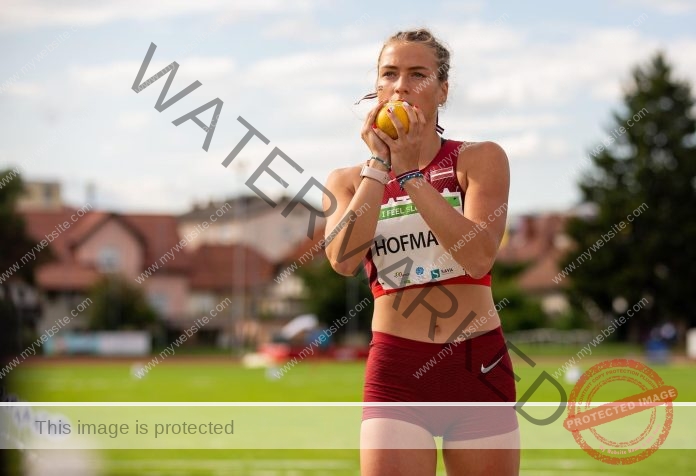 Anna Hofmane Latvian track and field athlete Anna Hofmane, in red sports gear, holds a shot by her chin on a field; houses in background.