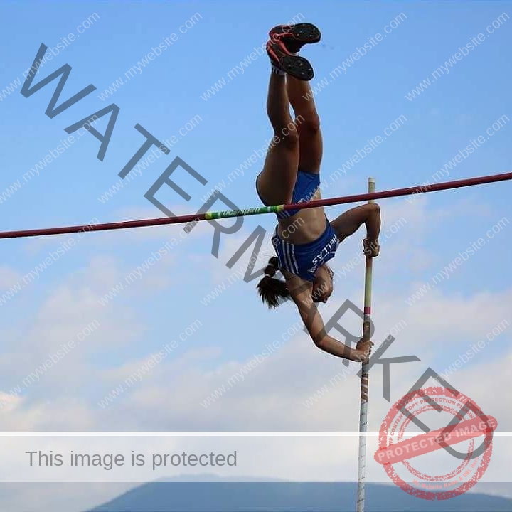 ariadni-adamopoulou-greece-ariadni_adam-08983 Ariadni Adamopoulou, pole vaulter from Greece and Oklahoma State clears the bar mid-air with one hand, blue sky and mountains behind.