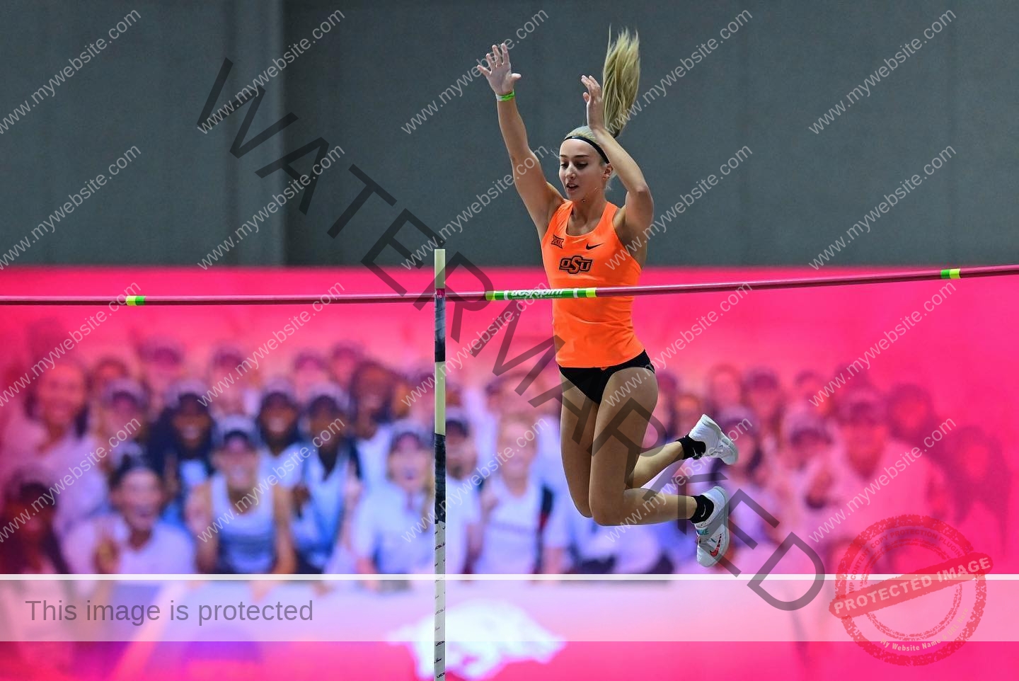 ariadni-adamopoulou-greece-ariadni_adam-08985 Ariadni Adamopoulou, pole vaulter from Greece and Oklahoma State, in orange OSU gear mid-air indoors, arms raised, pink backdrop.