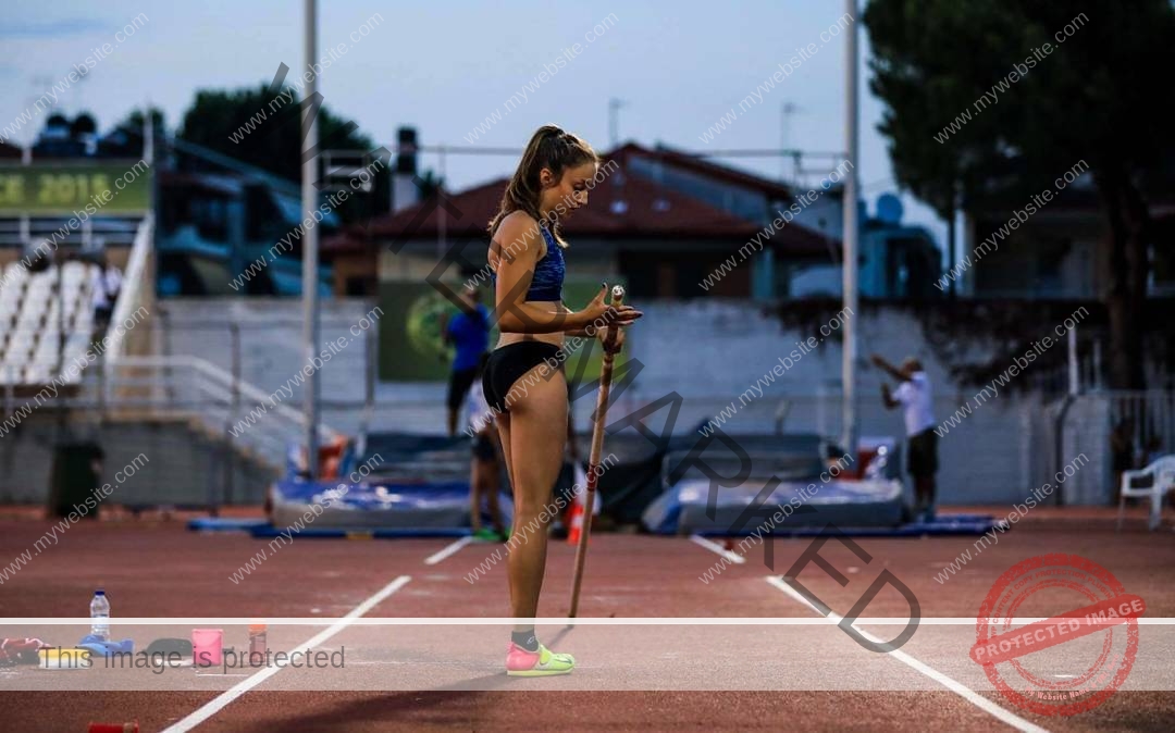 ariadni-adamopoulou-greece-ariadni_adam-08994 Ariadni Adamopoulou, pole vaulter from Greece and Oklahoma State, stands on a dusk track, pole ready, with gear and athletes nearby.
