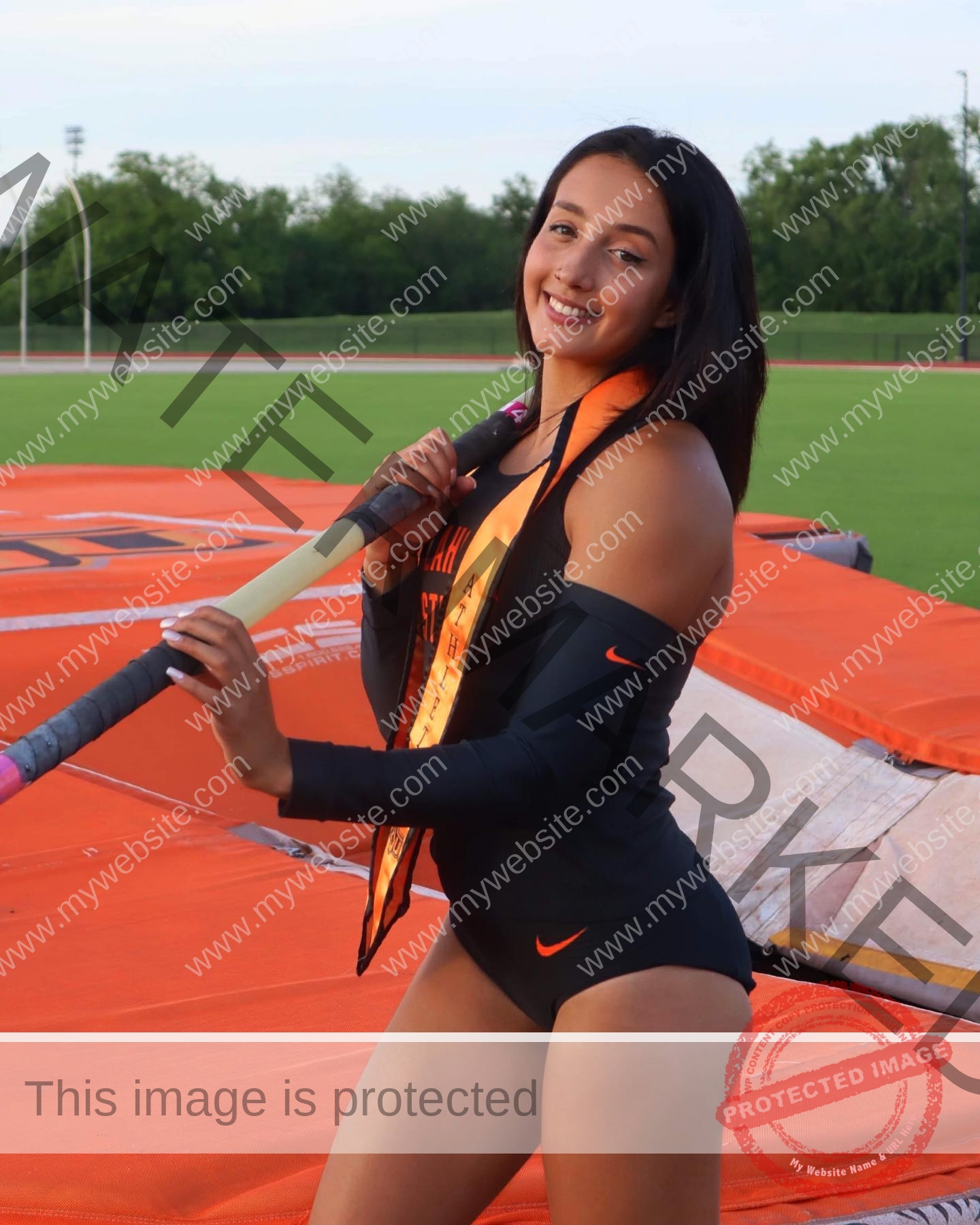 ariadni-adamopoulou-greece-ariadni_adam-09030 Ariadni Adamopoulou, pole vaulter from Greece, stands on a track in black sportswear, holding a pole and smiling; mats and grass behind.