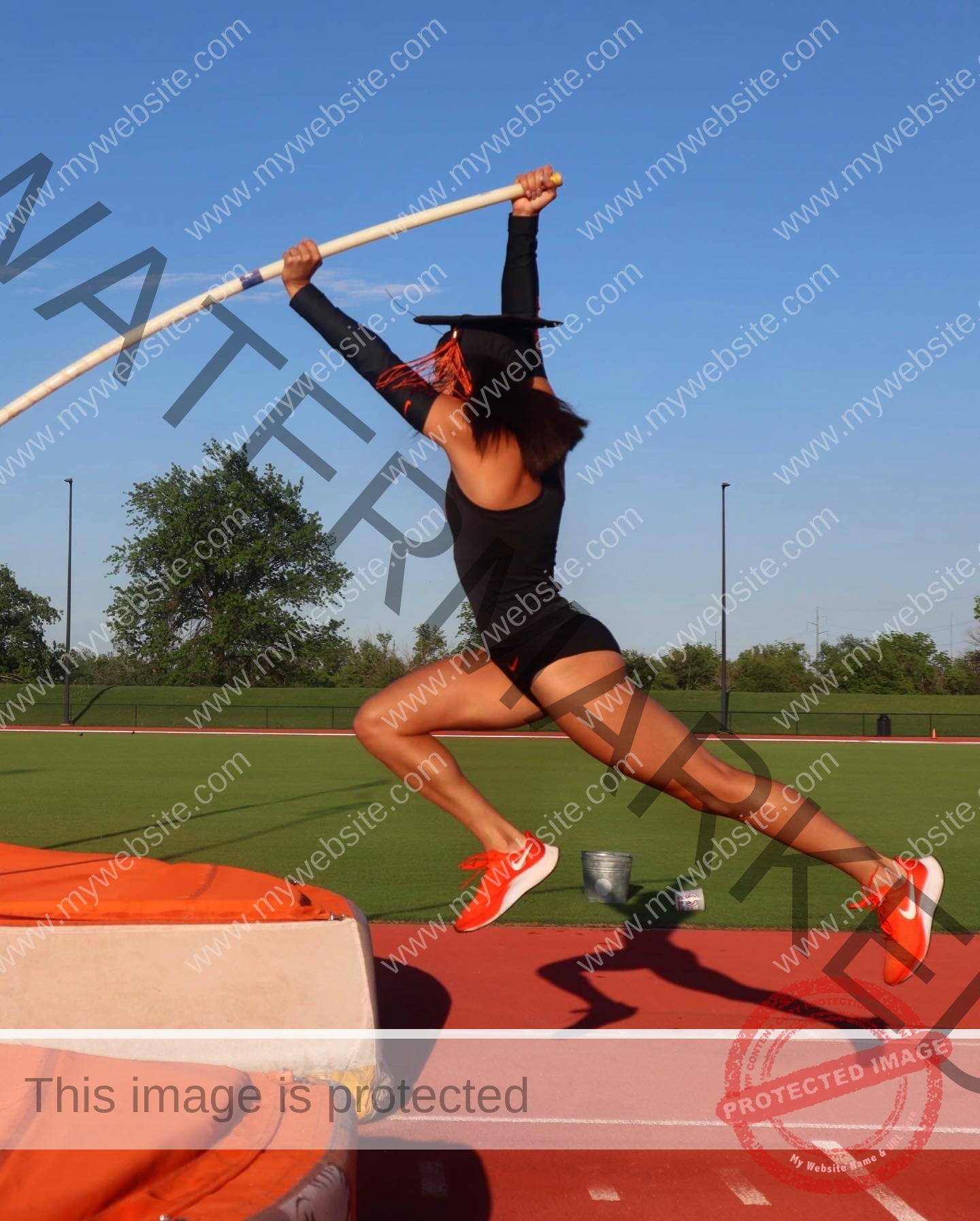 ariadni-adamopoulou-greece-ariadni_adam-09031 Ariadni Adamopoulou, pole vaulter from Greece, wearing athletic gear and a graduation cap vaults over a track with grass and blue sky.