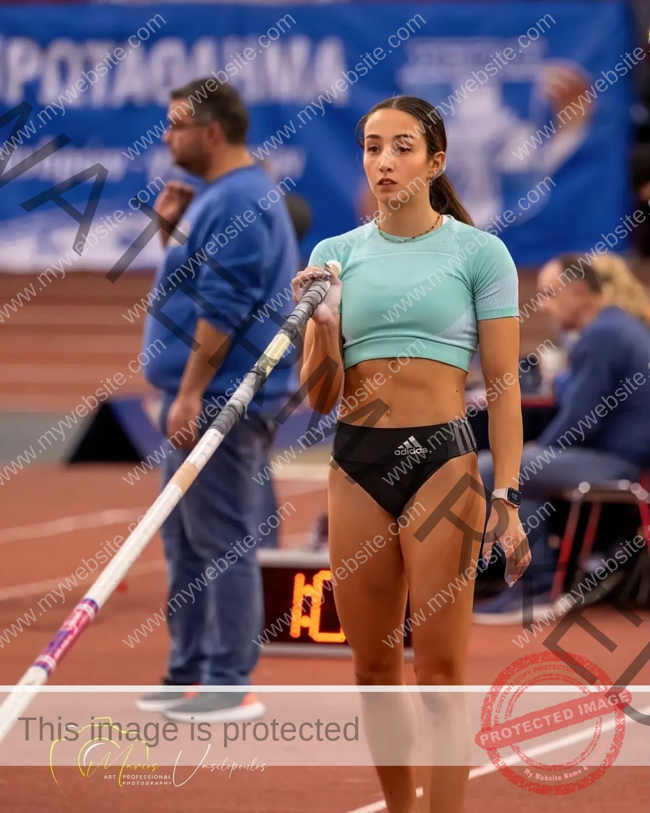 ariadni-adamopoulou-greece-ariadni_adam-09050 Ariadni Adamopoulou, pole vaulter from Greece, in a light blue crop top and black shorts holds a pole at an indoor track event.