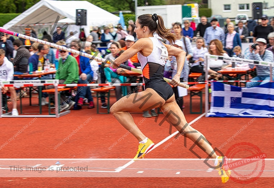 ariadni-adamopoulou-greece-ariadni_adam-09079 Ariadni Adamopoulou, pole vaulter from Greece and Oklahoma State, runs on a red track holding a pole, preparing to jump; spectators and Greek flag in background.