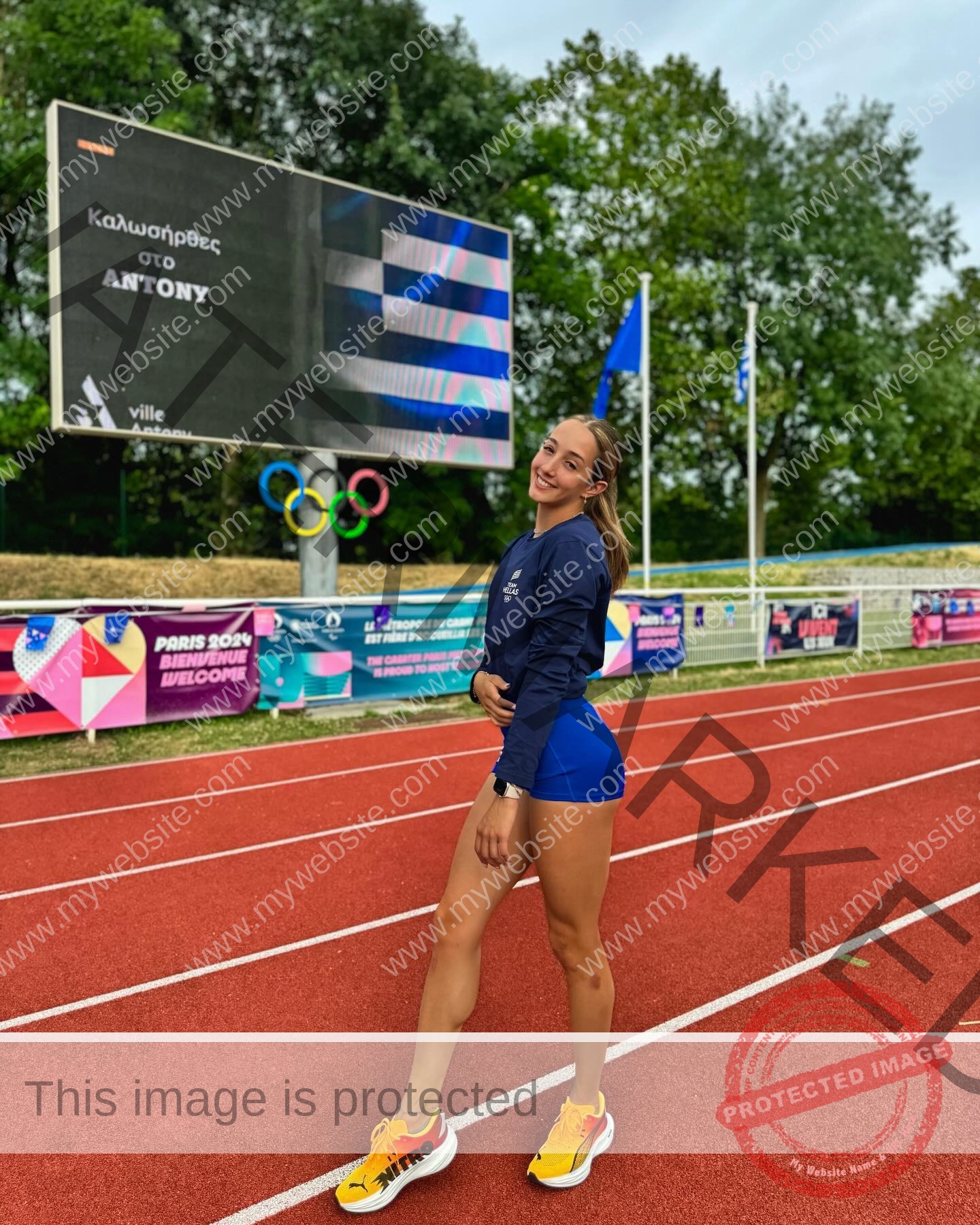 ariadni-adamopoulou-greece-ariadni_adam-09083 Ariadni Adamopoulou, pole vaulter from Greece and Oklahoma State, stands smiling on a red track with banners, flags, and trees behind.