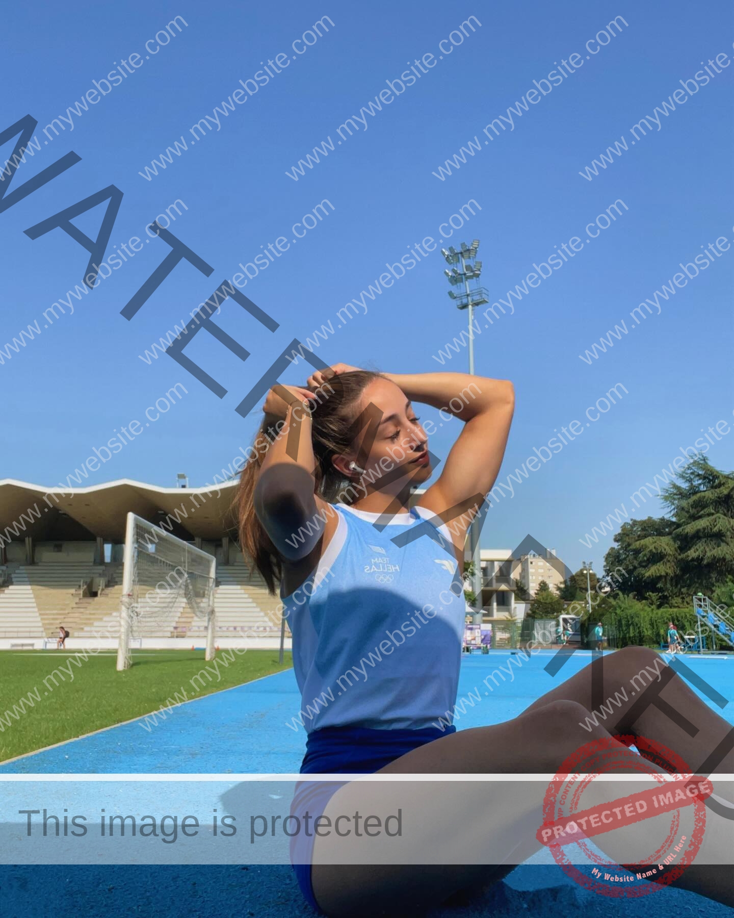 ariadni-adamopoulou-greece-ariadni_adam-09084 Ariadni Adamopoulou, pole vaulter from Greece and Oklahoma State, sits on a blue track tying her hair; stadium seats and goal behind.