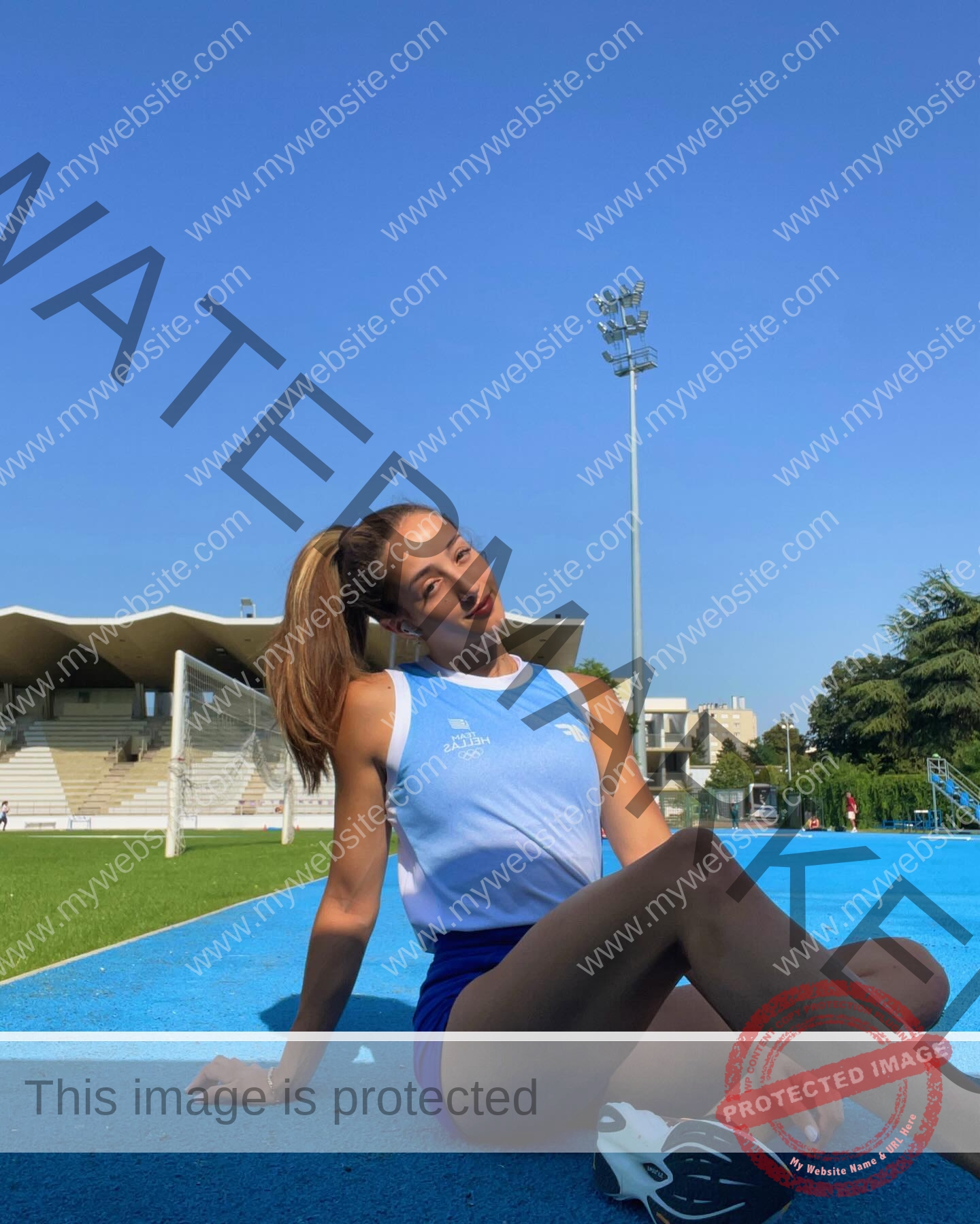 ariadni-adamopoulou-greece-ariadni_adam-09085 Ariadni Adamopoulou, pole vaulter from Greece and Oklahoma State, sits on a blue track in light blue athletic gear, smiling.