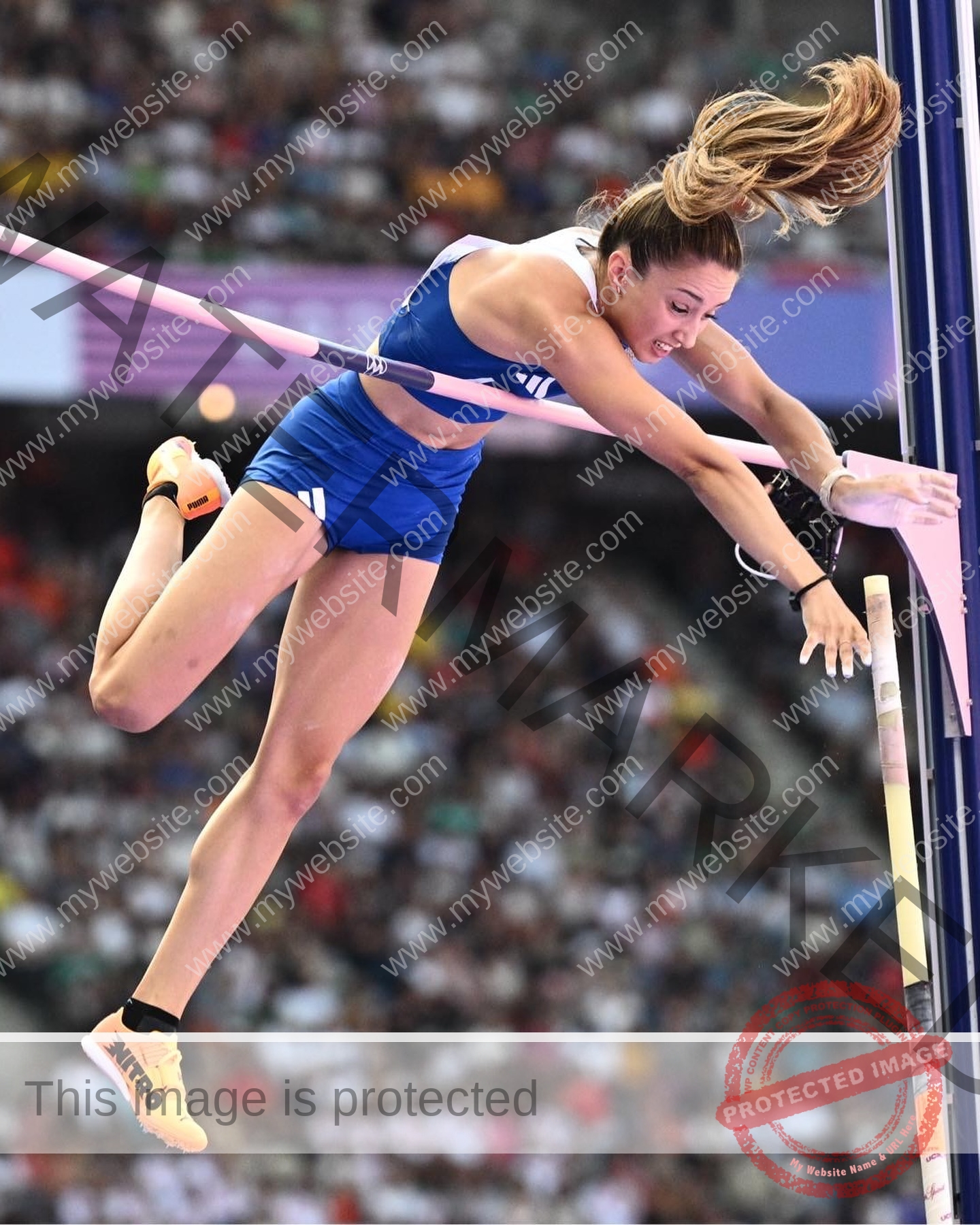 ariadni-adamopoulou-greece-ariadni_adam-09095 Ariadni Adamopoulou, pole vaulter from Greece and Oklahoma State, in mid-air wearing blue, clears the bar with stadium crowd blurred.