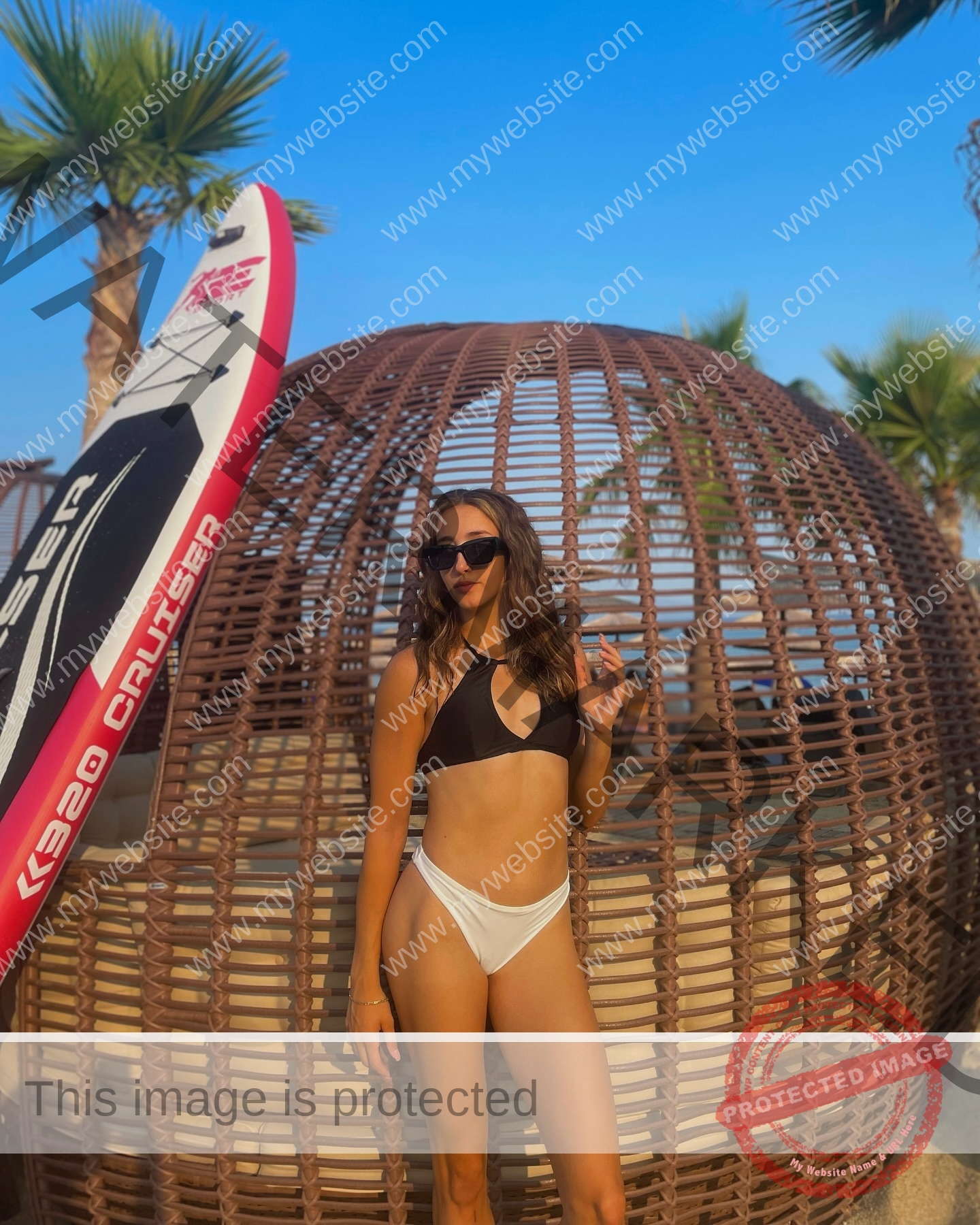 ariadni-adamopoulou-greece-ariadni_adam-09101 Ariadni Adamopoulou, pole vaulter from Greece and Oklahoma State, wearing a bikini stands by a wicker dome, surfboard at her side.