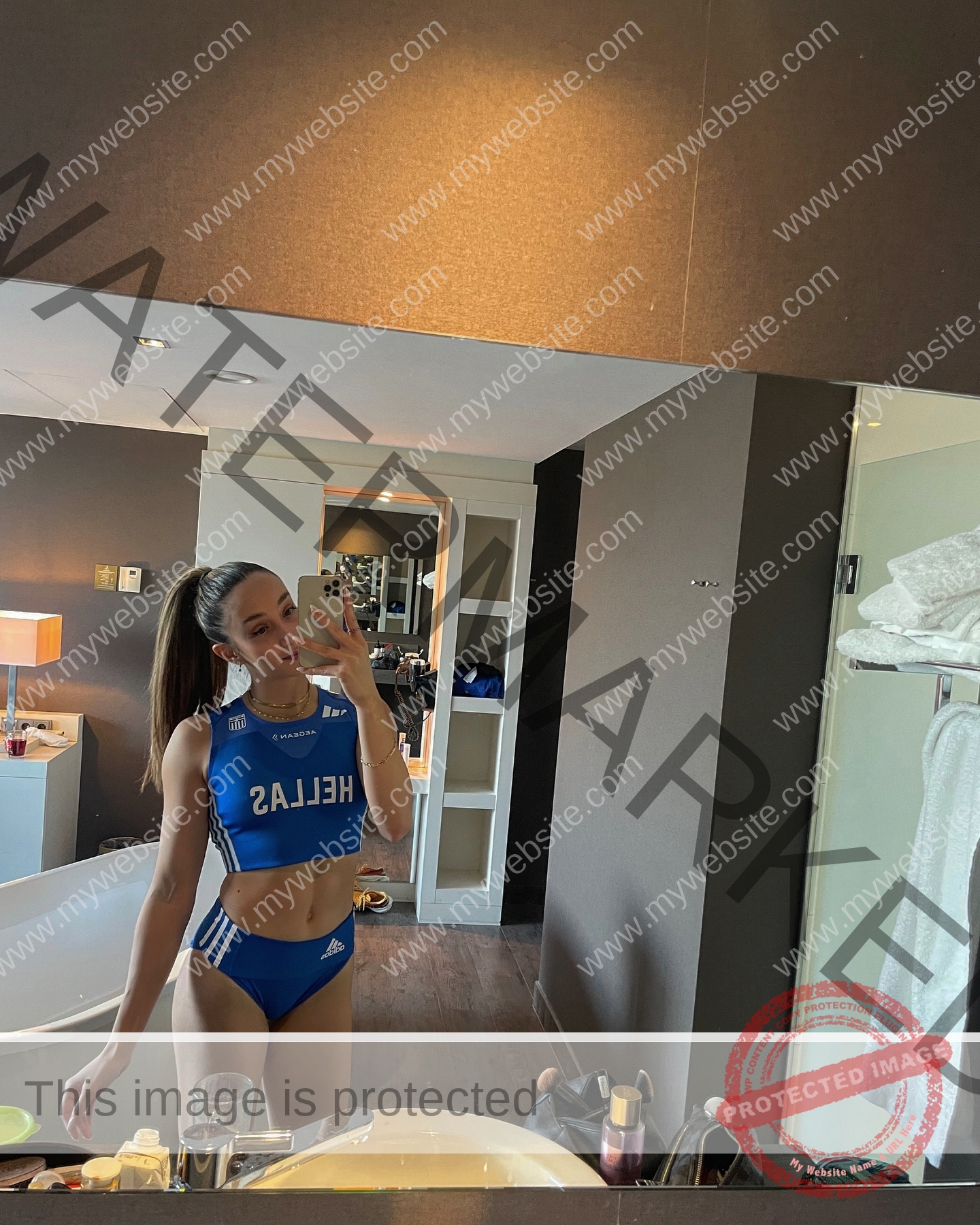 ariadni-adamopoulou-greece-ariadni_adam-09131 Basssant Hemida, track star from Egypt, in a blue HELLAS sports outfit takes a mirror selfie in a modern bathroom, toiletries visible.