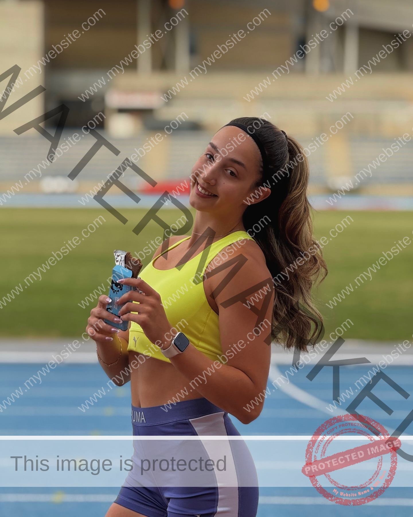 ariadni-adamopoulou-greece-ariadni_adam-09140 Ariadni Adamopoulou, pole vaulter from Greece and Oklahoma State, smiles in athletic wear on a blue track, holding a snack bar.