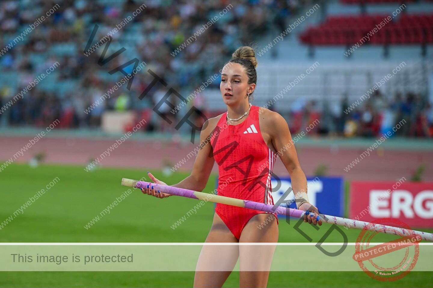 ariadni-adamopoulou-greece-ariadni_adam-09158 Ariadni Adamopoulou, pole vaulter from Greece and Oklahoma State, in red gear holds her pole on a stadium track, crowd blurred.