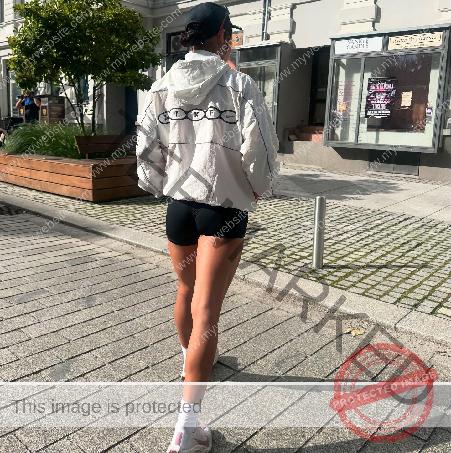 arie-aoki-japan-aokiarie_-x-09177 Arie Aoki, track star from Japan, walks on a sunny city sidewalk in a white Nike jacket, black shorts, cap, and white socks.