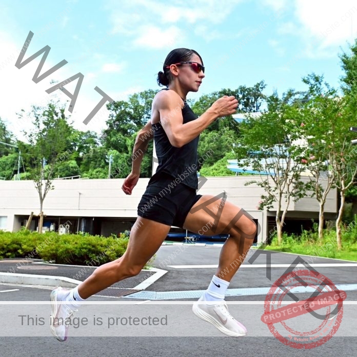 Arie Aoki, track star from Japan, wearing sunglasses, a black tank top, and shorts is mid-air running outdoors on pavement.