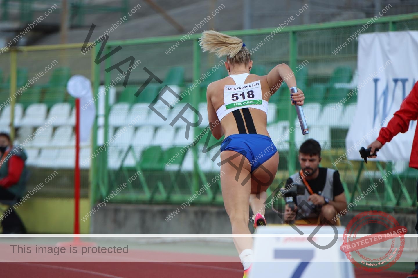 birna-kristjansdottir-iceland-birnakristink-10911 Birna-Kristjansdottir, track athlete from Iceland, sprints in blue and white with baton, number 518; empty seats and fence behind.