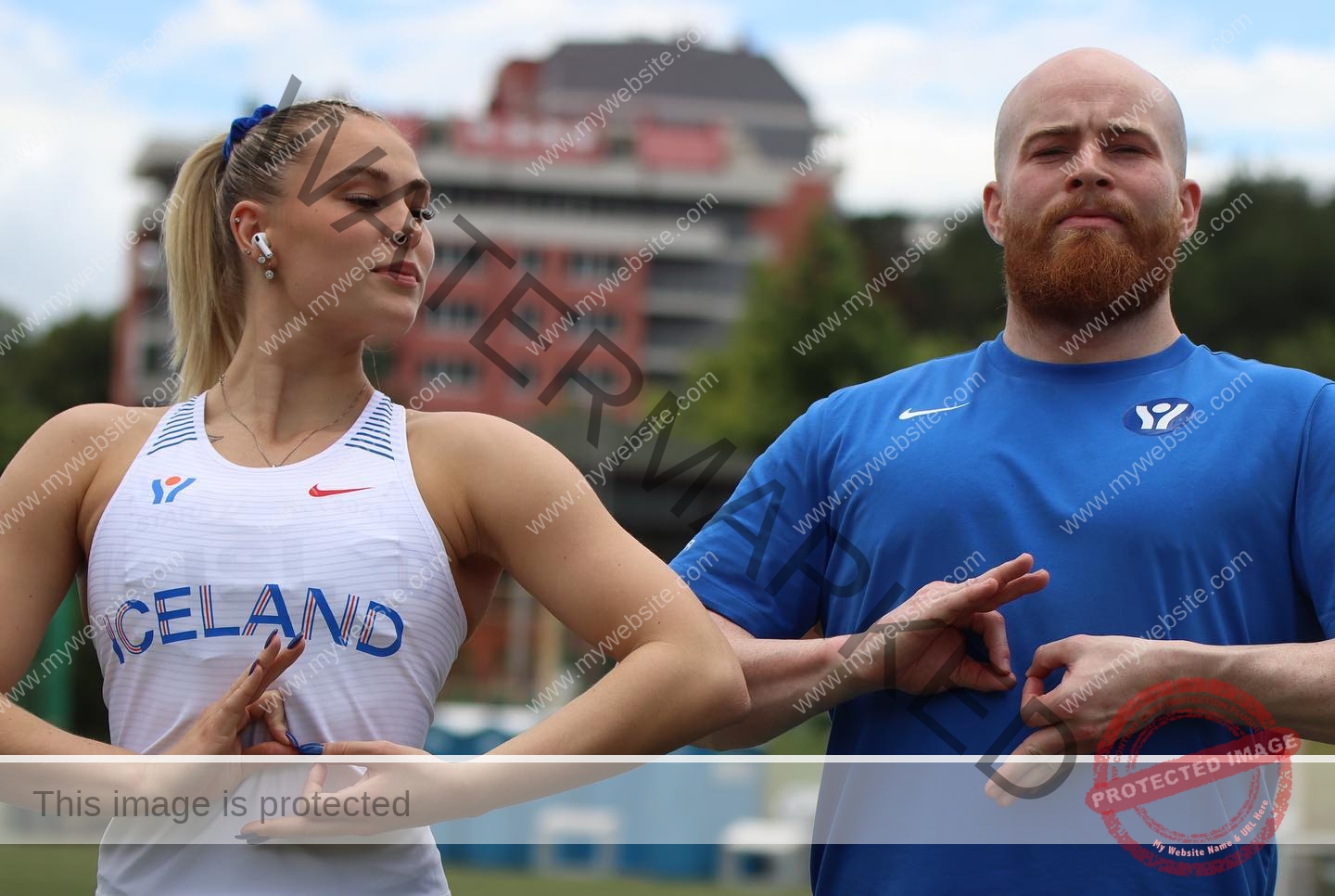 birna-kristjansdottir-iceland-birnakristink-10915 Birna-Kristjansdottir, track athlete from Iceland, and a man in blue make matching hand signs outdoors near a red building and trees.