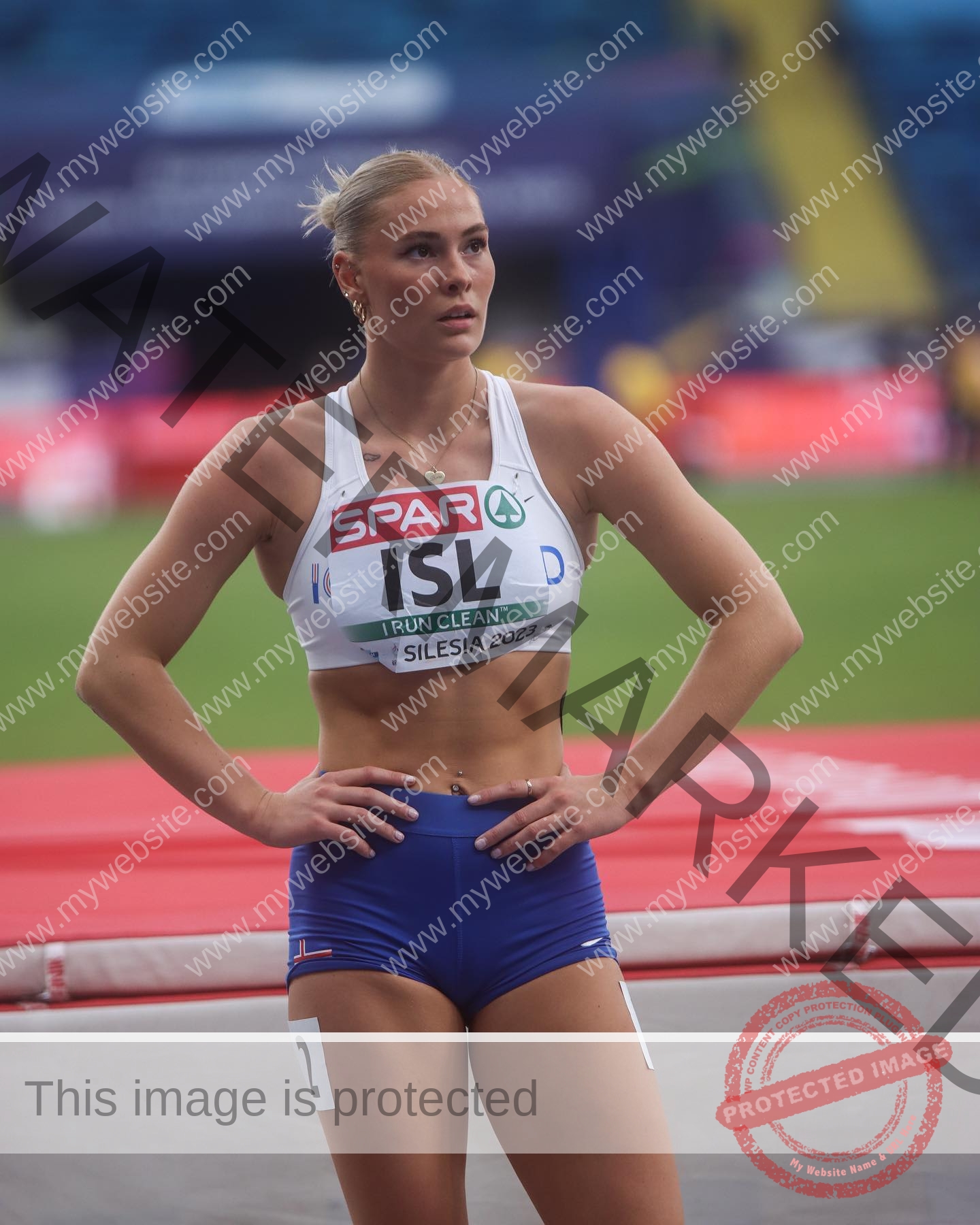 birna-kristjansdottir-iceland-birnakristink-10934 Birna-Kristjansdottir, track athlete from Iceland stands with hands on hips on a track in ISL jersey, focused, stadium blurred.
