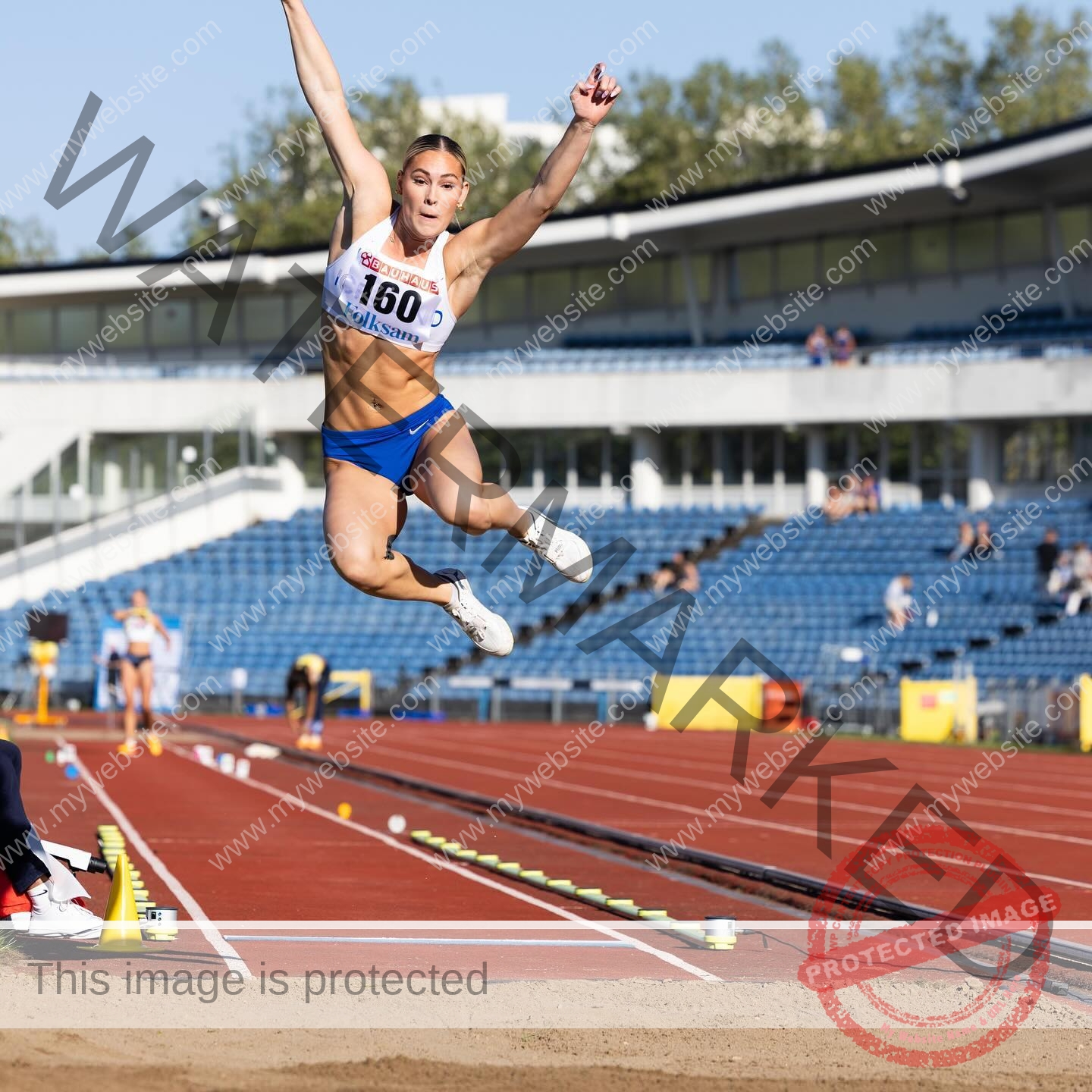 birna-kristjansdottir-iceland-birnakristink-10961 Birna-Kristjansdottir, track athlete from Iceland, is in mid-air during the long jump at an outdoor stadium with blue seats and trees.