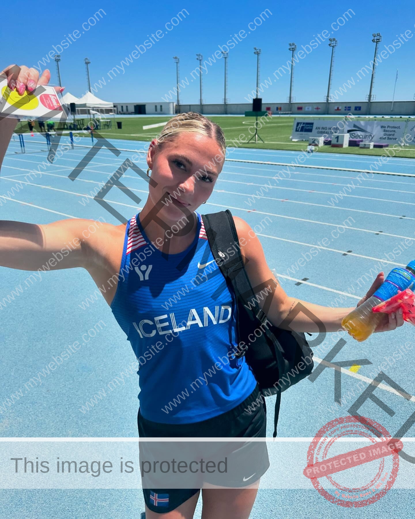 birna-kristjansdottir-iceland-birnakristink-10991 Birna-Kristjansdottir, track athlete from Iceland, smiles on a blue track in her blue jersey, holding a drink and paper with a backpack.