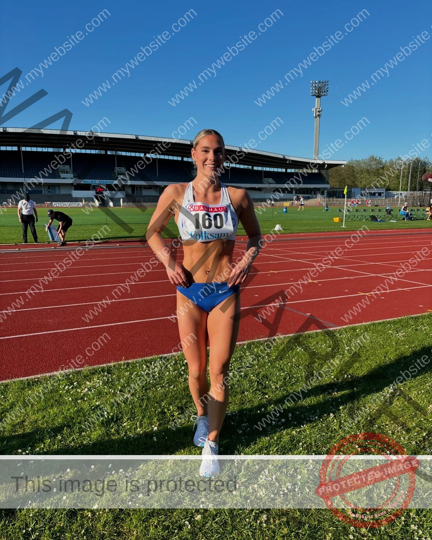 birna-kristjansdottir-iceland-birnakristink-10999 Birna-Kristjansdottir, track athlete from Iceland, stands smiling on grass by a track in sports bra, blue shorts, bib 160.