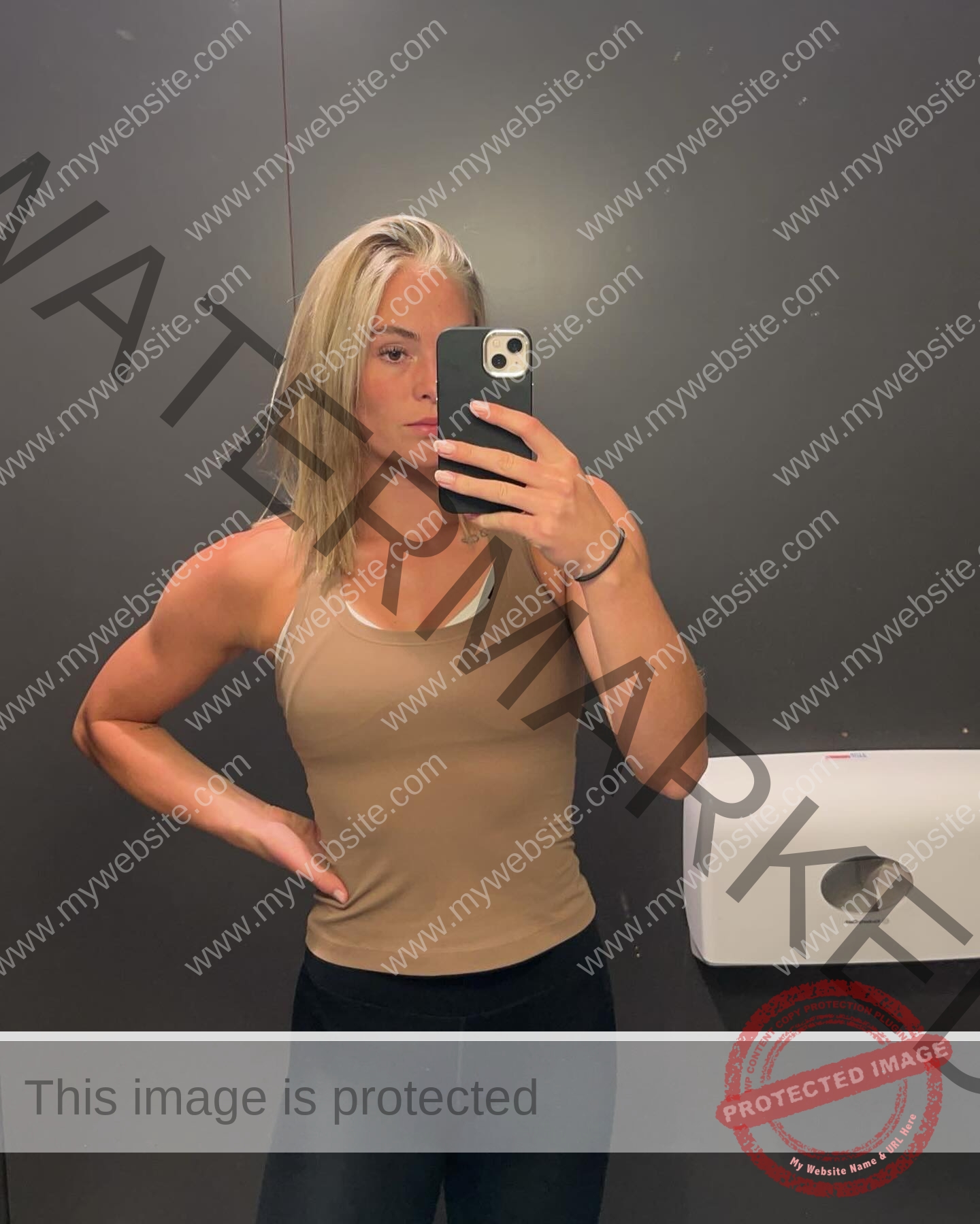 birna-kristjansdottir-iceland-birnakristink-11012 Birna-Kristjansdottir, track athlete from Iceland, takes a mirror selfie in a tan tank top and black pants by a wall dispenser.