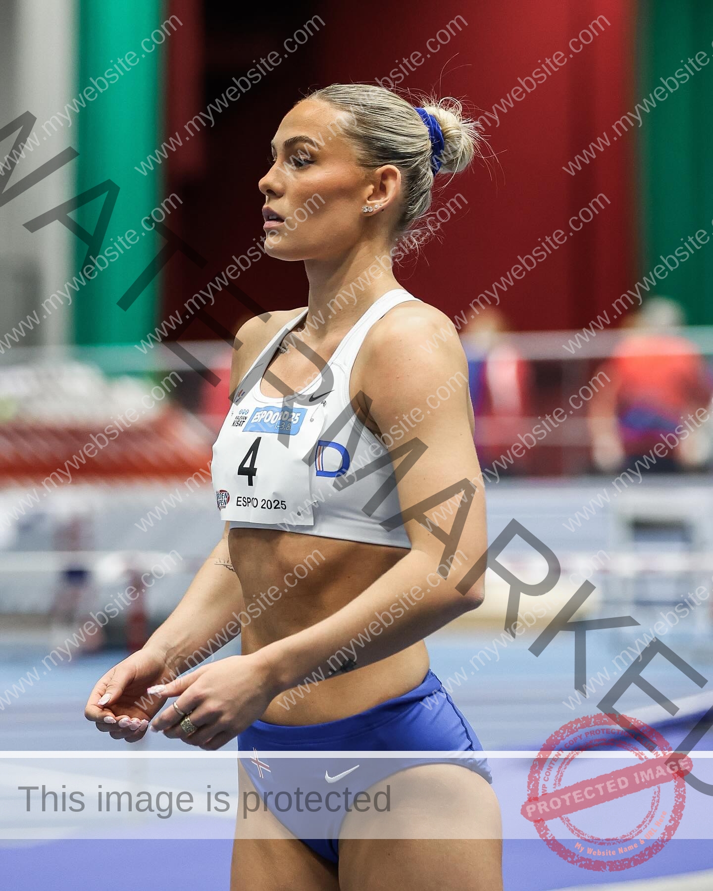birna-kristjansdottir-iceland-birnakristink-11026 Birna-Kristjansdottir, track athlete from Iceland, stands on an indoor track in a white crop top and blue shorts, focused, bib number 4.