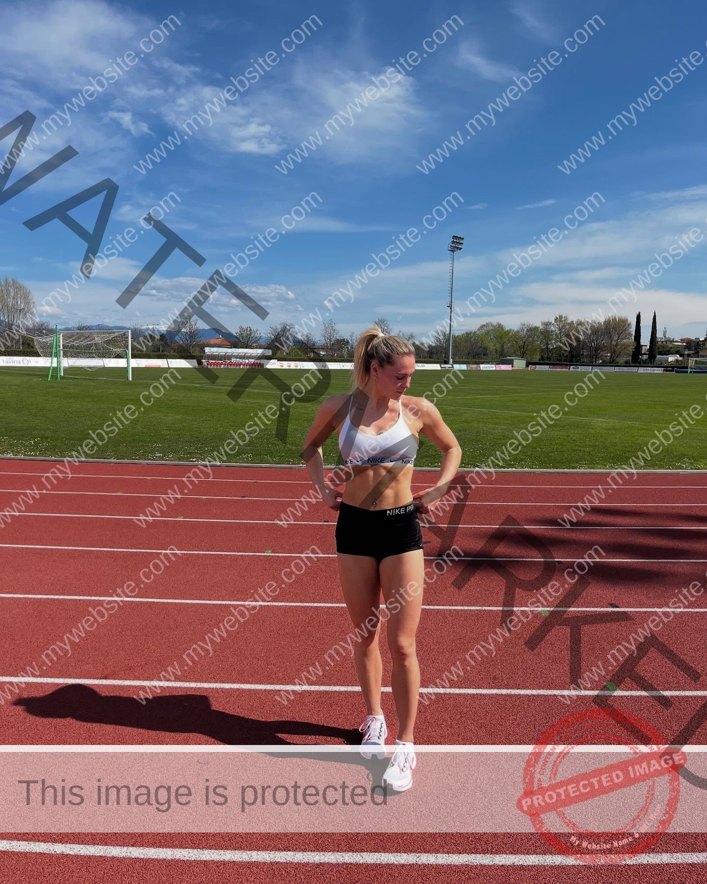 birna-kristjansdottir-iceland-birnakristink-11041 Birna-Kristjansdottir, track athlete from Iceland, in athletic wear stands on a red track under blue sky, hands on hips, field gear behind.