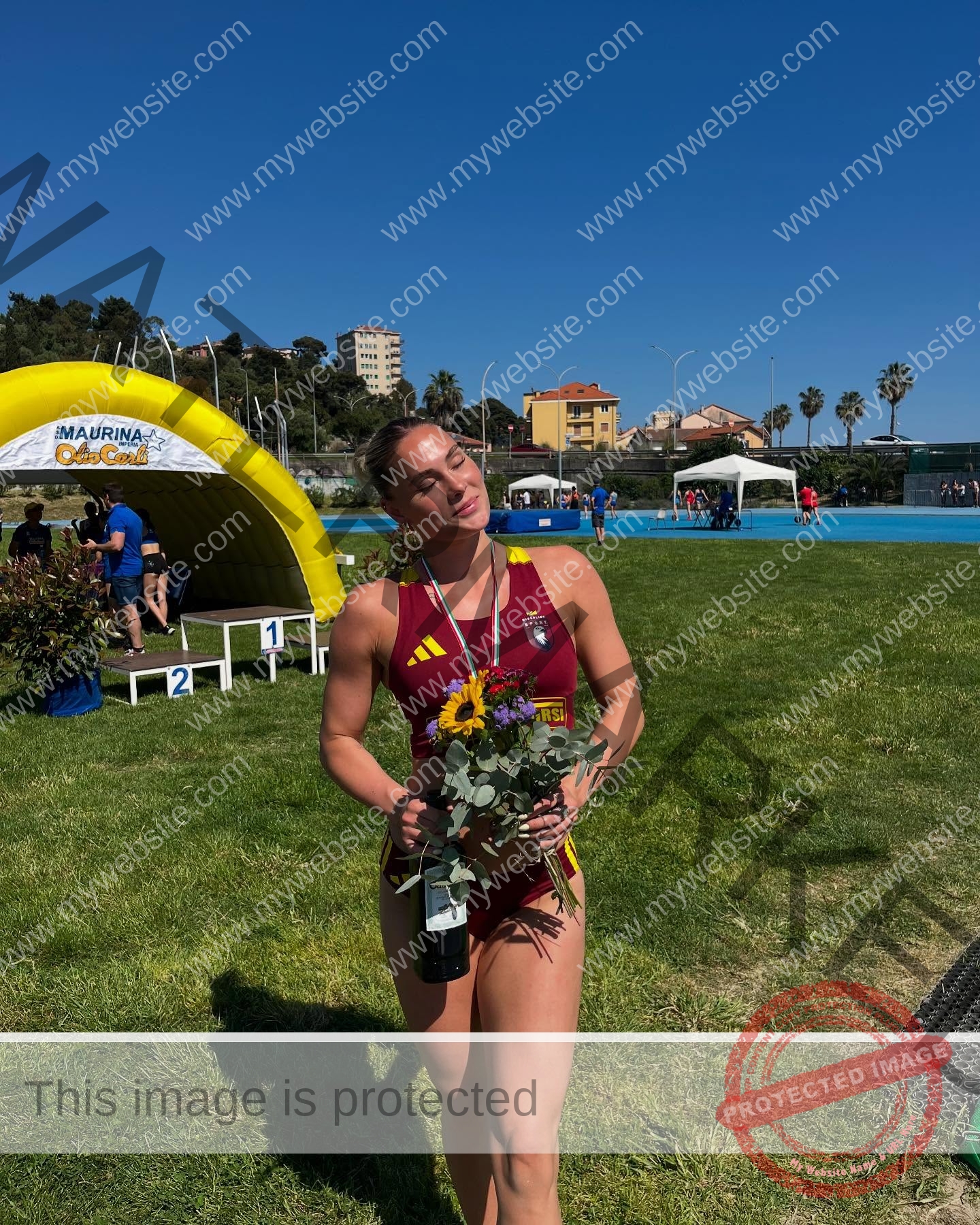 birna-kristjansdottir-iceland-birnakristink-11050 Birna-Kristjansdottir, track athlete from Iceland, in red stands on grass with bouquet, medal, and bottle, smiling near a yellow arch.